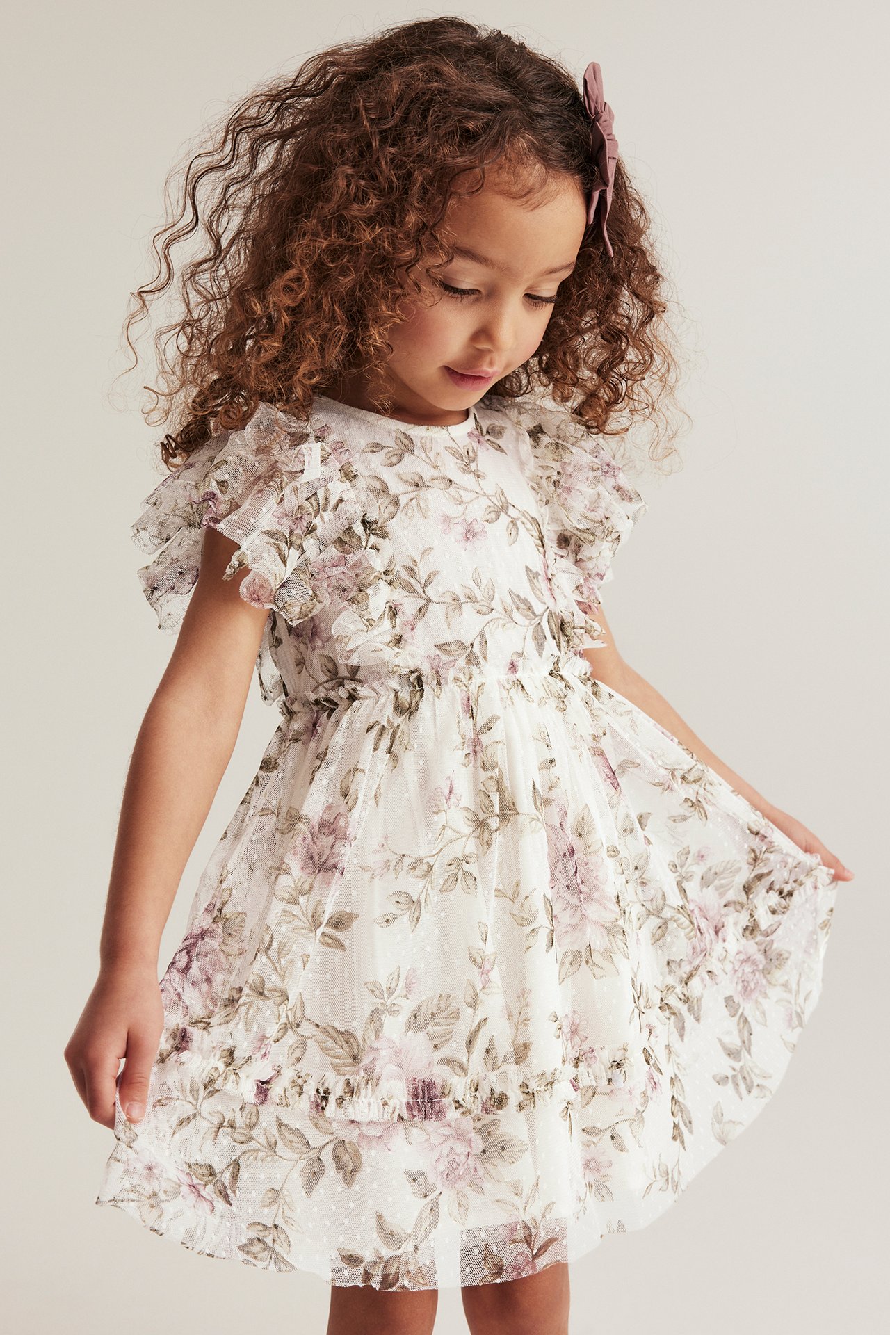 Front view of a kid wearing an off-white floral mesh dress with ruffle sleeves and a pink hair bow.