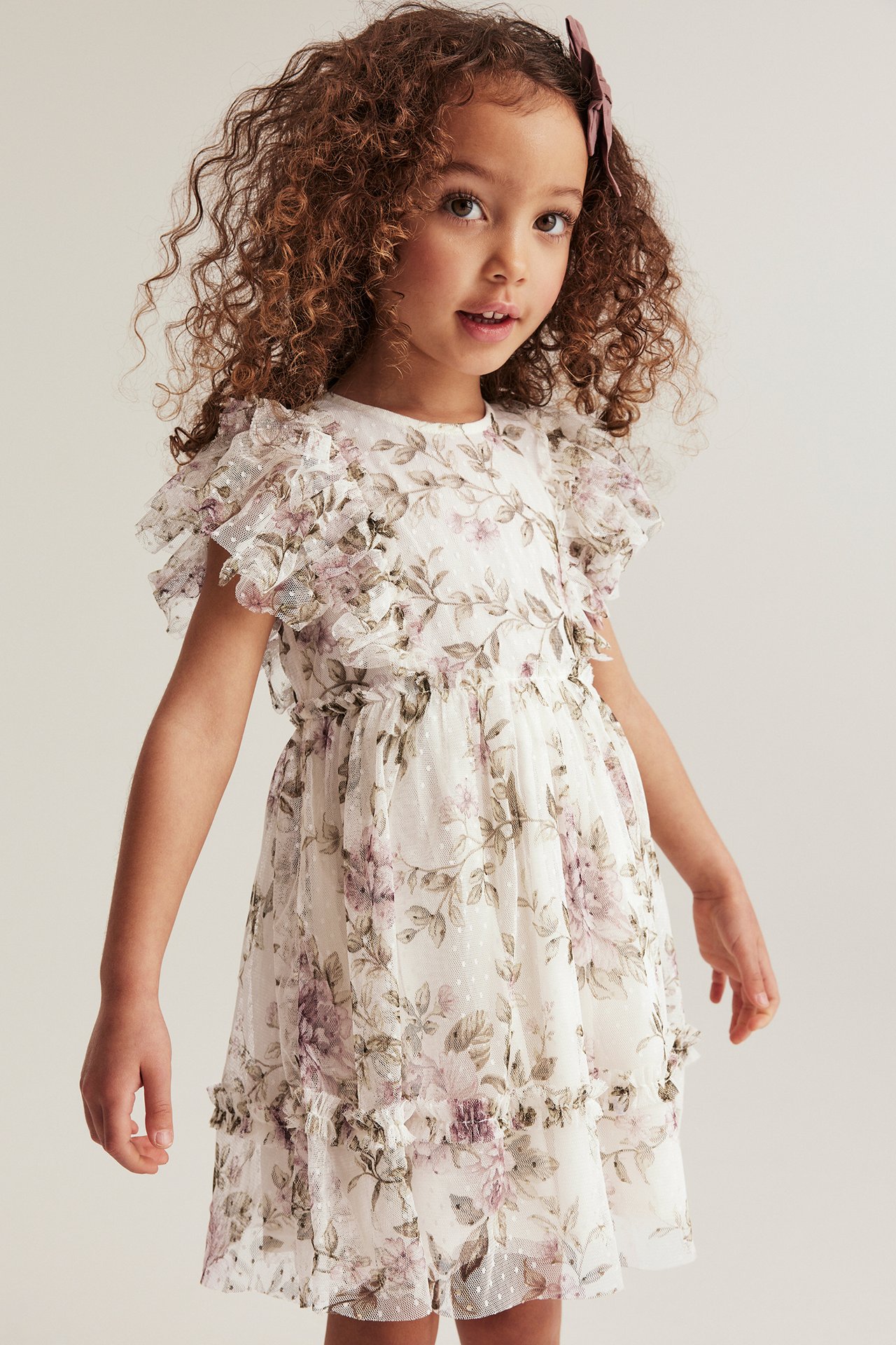 Front view of a kid girl wearing an off-white floral mesh dress with ruffles and a purple hair clip.