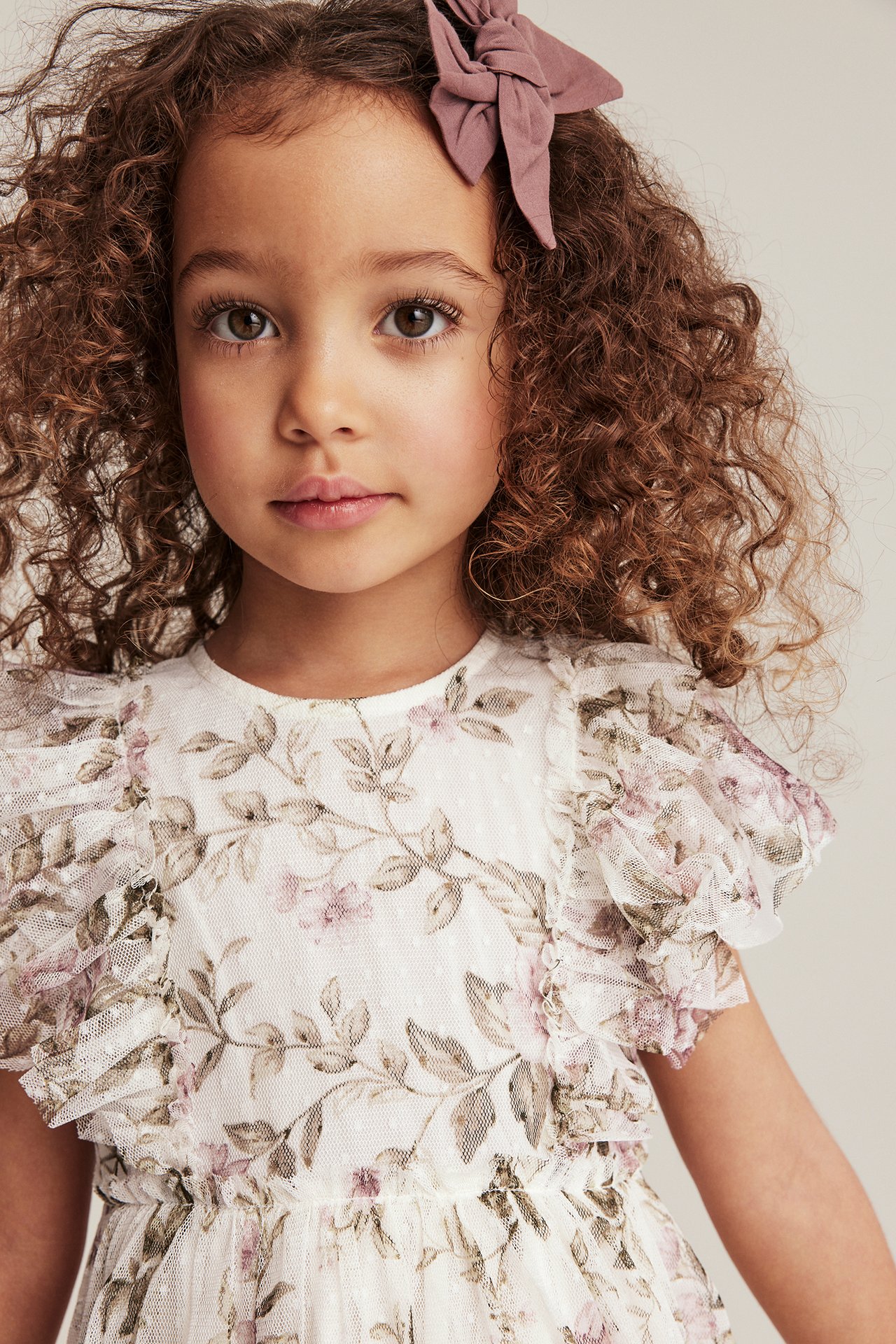 Close-up of a kid in an off-white floral mesh dress with ruffle sleeves and a pink bow.