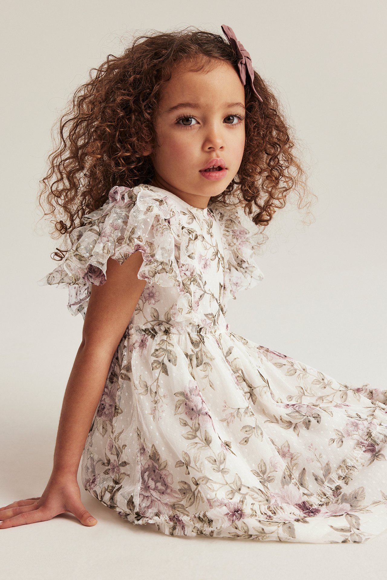 Front view of a kid in an off-white floral mesh dress with ruffle sleeves and a purple hair bow.