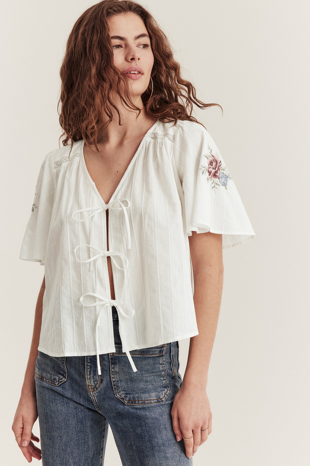 Front view: woman in offwhite woven cotton blouse with floral embroidery, tie-front, blue jeans.