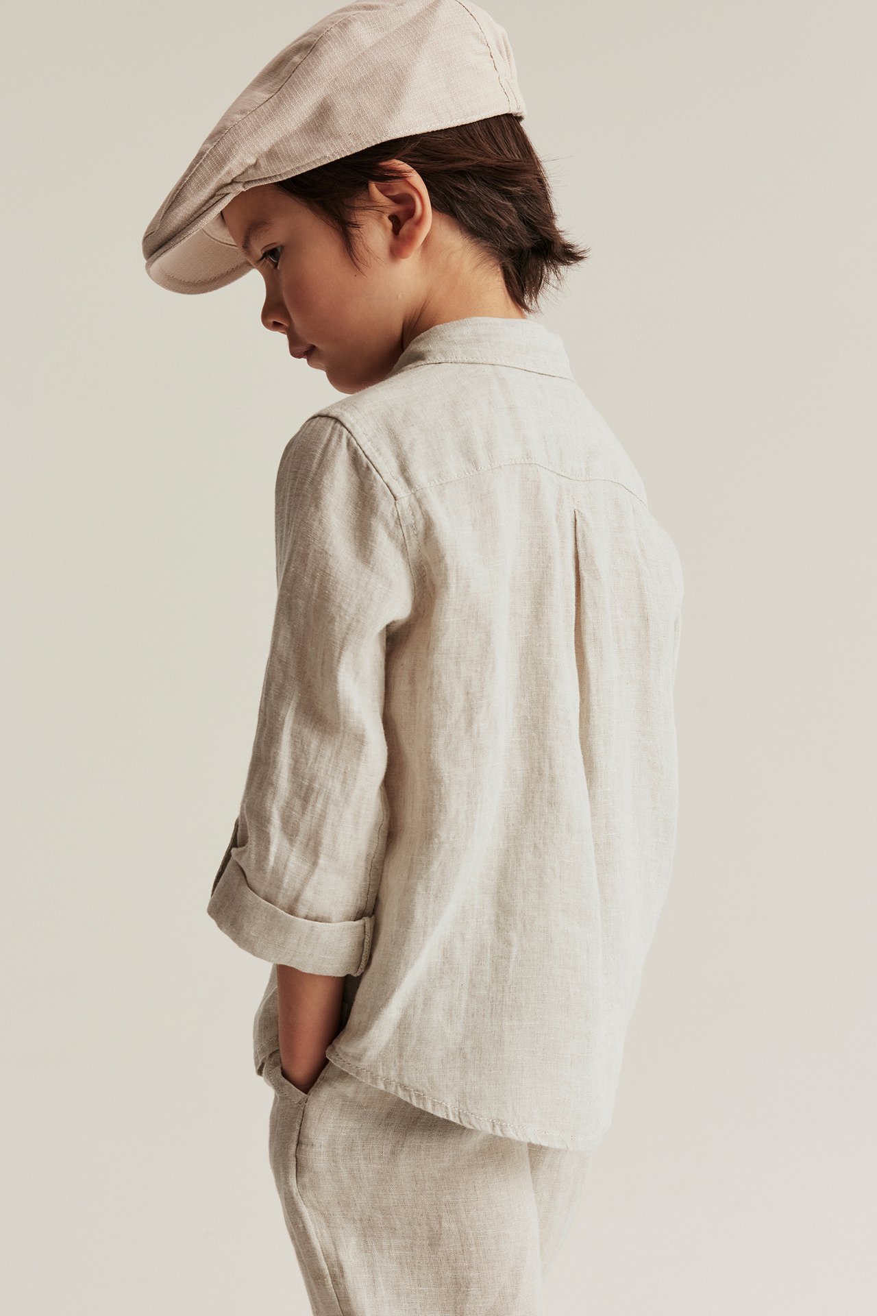 Side view of a kid wearing a light beige linen shirt with rolled-up sleeves, matching pants, and a flat cap.