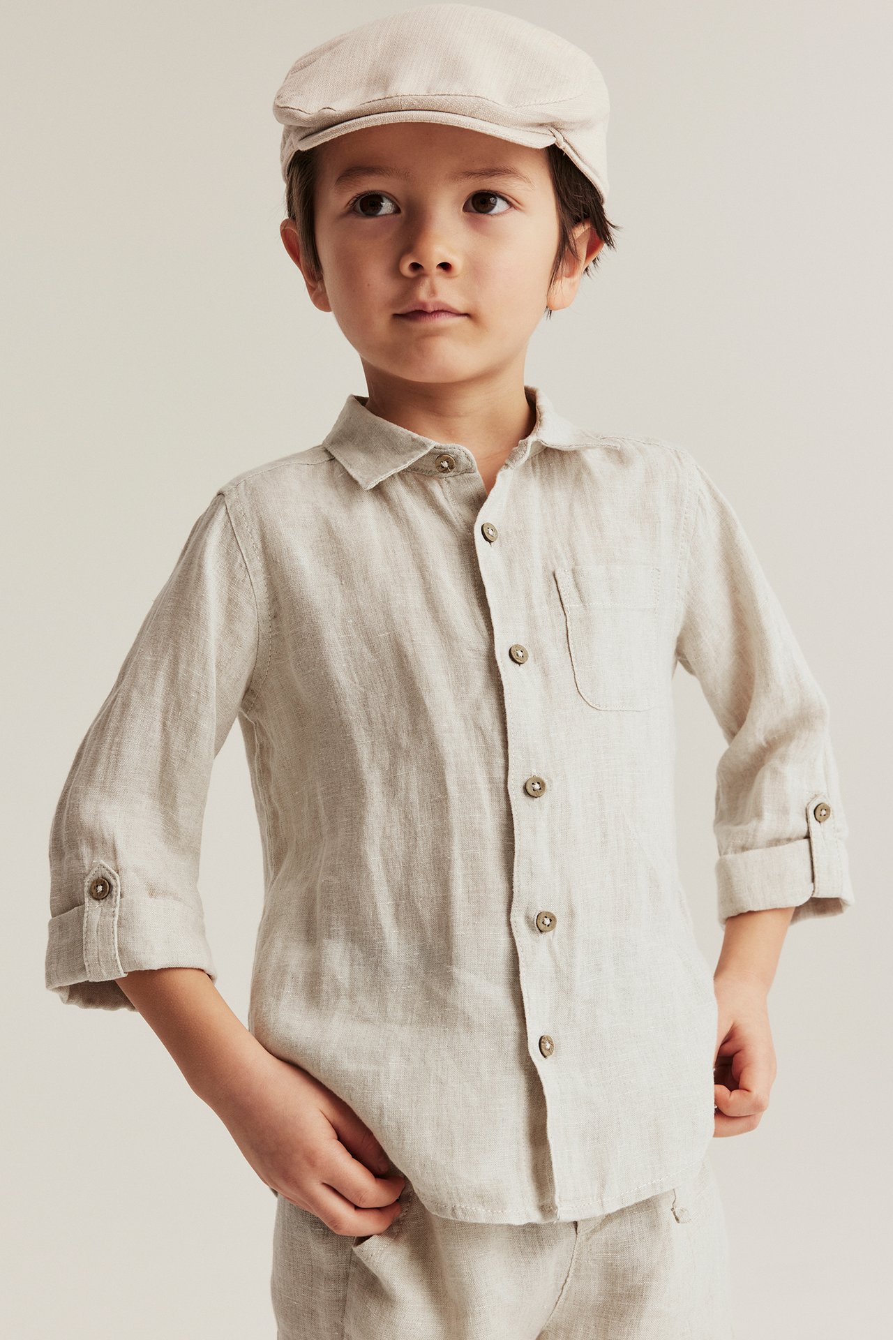 Front view of a kid in a light beige linen shirt with rolled sleeves, chest pocket, and matching flat cap.