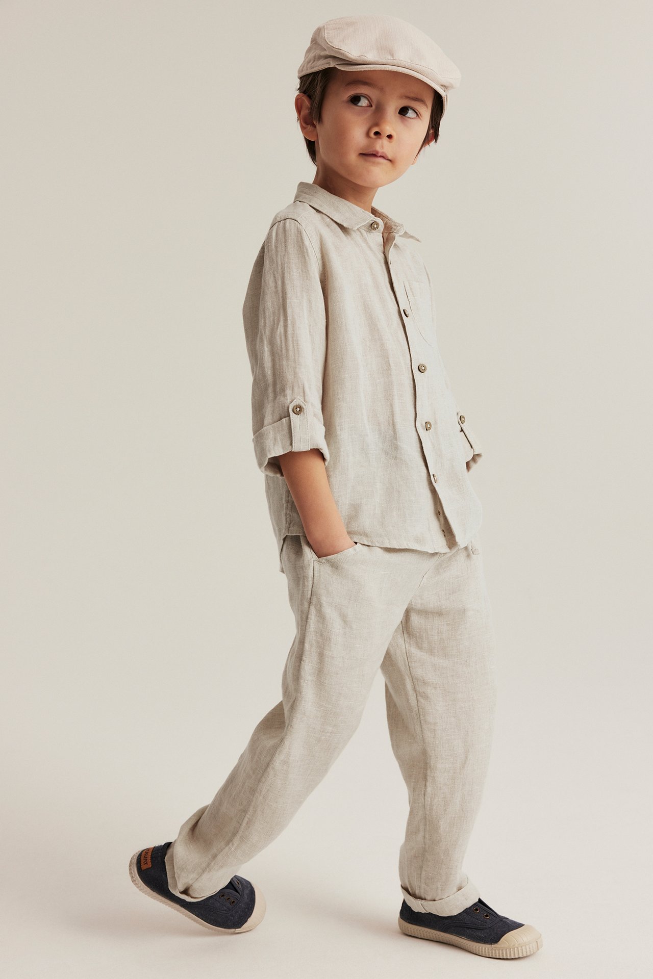 Kid wears a light beige linen shirt, matching trousers, a cap, and dark blue shoes. Front view.