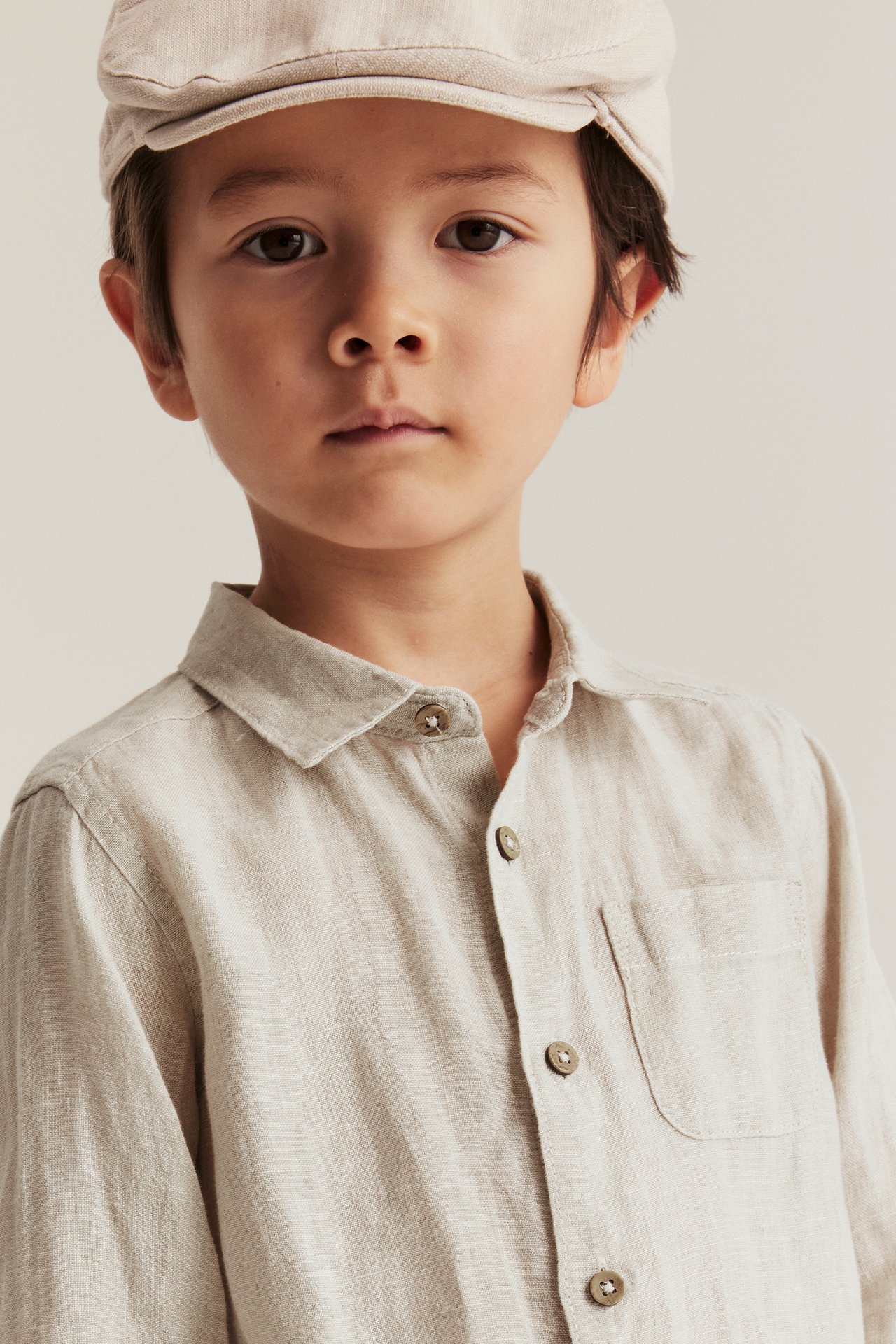 Close-up front view of a kid wearing a light beige linen shirt with a chest pocket and a matching cap.