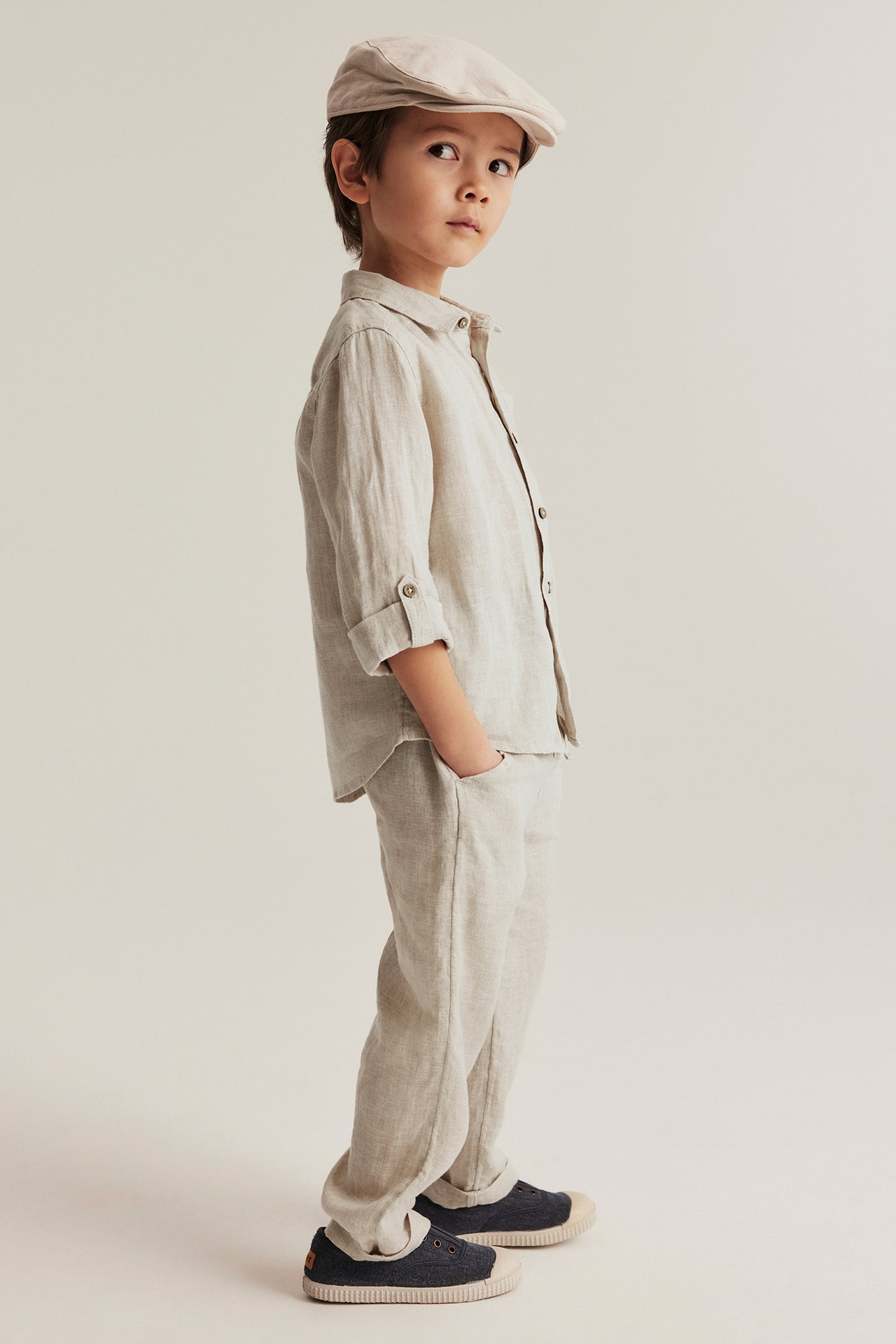 A kid wears a light beige linen shirt with rolled sleeves, matching trousers, a cap, and dark blue shoes.
