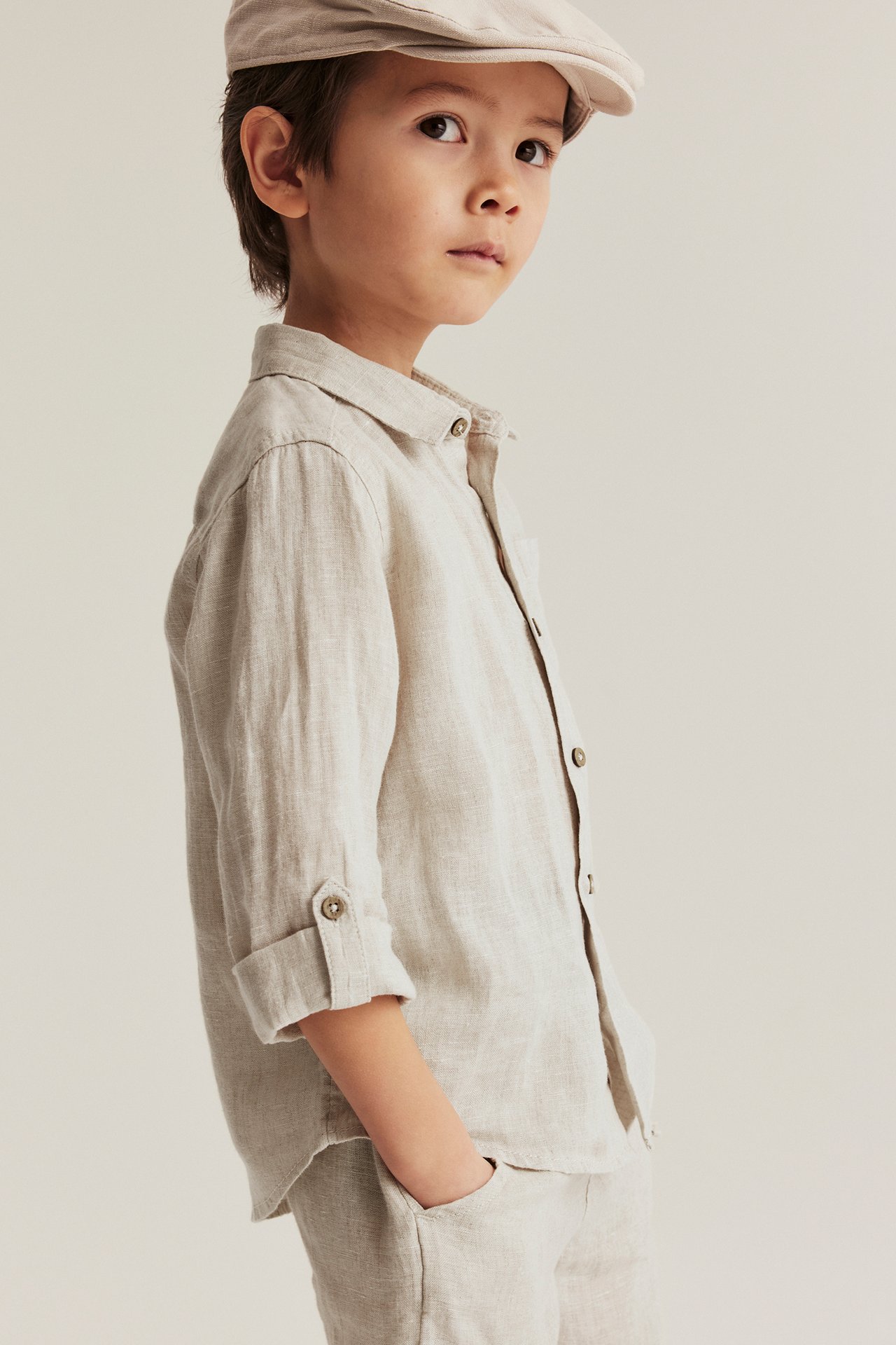 Side view of a kid in a light beige linen shirt with rolled sleeves, front buttons, and a matching cap.