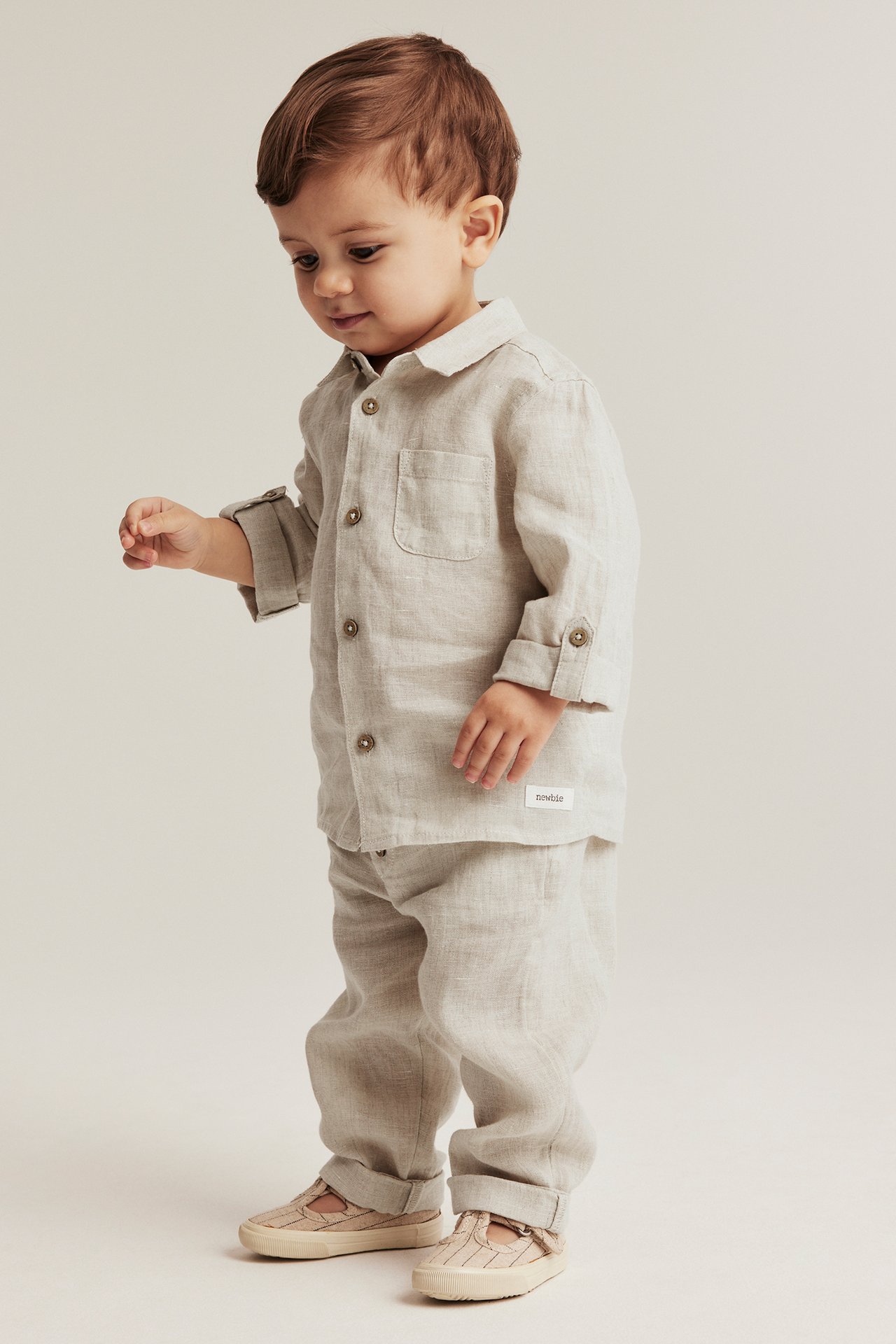 Seitenansicht eines Babys in hellbeiger Leinenhose mit Kordelzug, passendem Hemd und hellen T-Riemen-Schuhen.