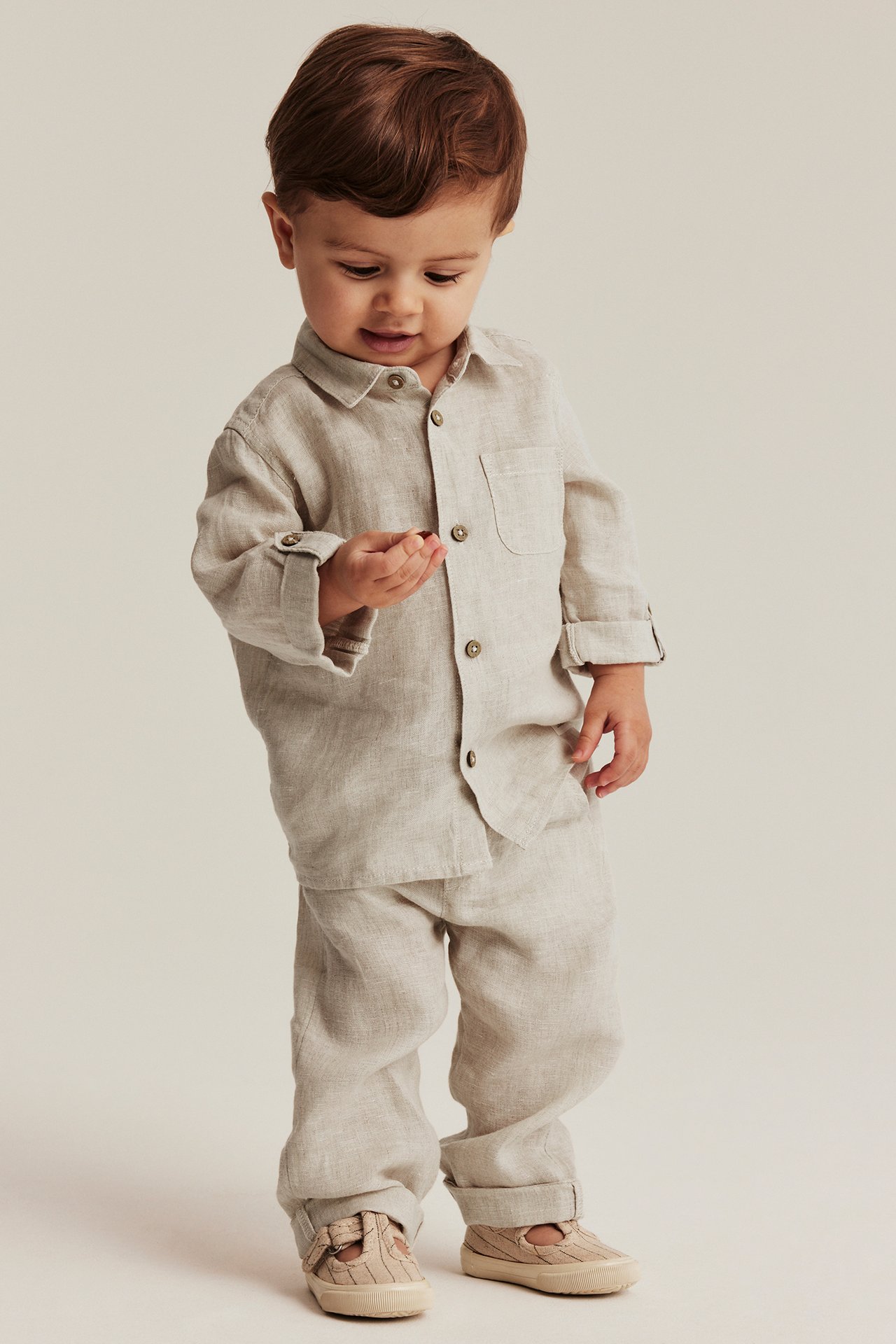Vorderansicht eines Babys in hellbeiger Leinenhose mit Kordelzug, passendem Leinenhemd und hellbeigen Schuhen.