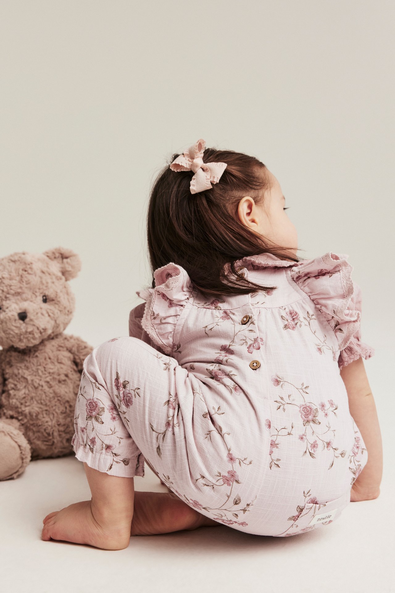 Back view: kid in pink floral cotton jumpsuit with ruffle sleeves, pink bow, next to teddy bear.