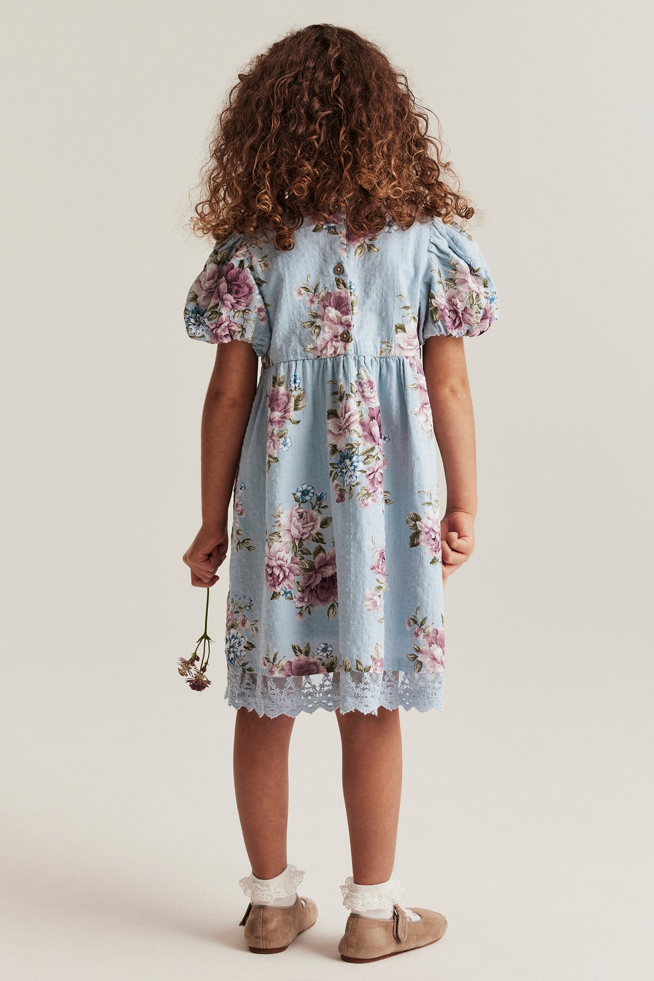 Rückenansicht eines Kindes: hellblaues geblümtes Baumwollkleid, Puffärmel, Knöpfe, Spitzenbesatz, beige Schuhe, weiße Socken.