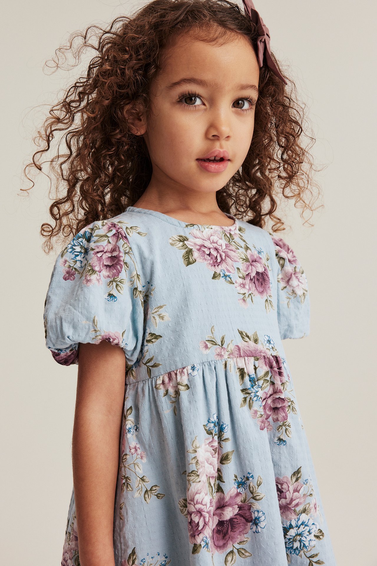 Nahaufnahme von vorne eines Kindes in einem hellblauen geblümten Baumwollkleid mit Ballonärmeln und lila Haarspange.