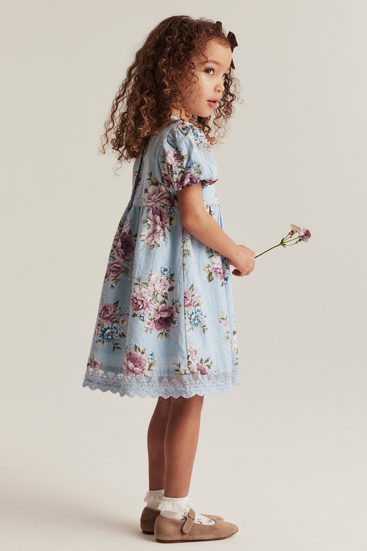 Seitenansicht eines Kindes in hellblauem geblümtem Baumwollkleid, weißen Spitzensocken, beigen Schuhen.
