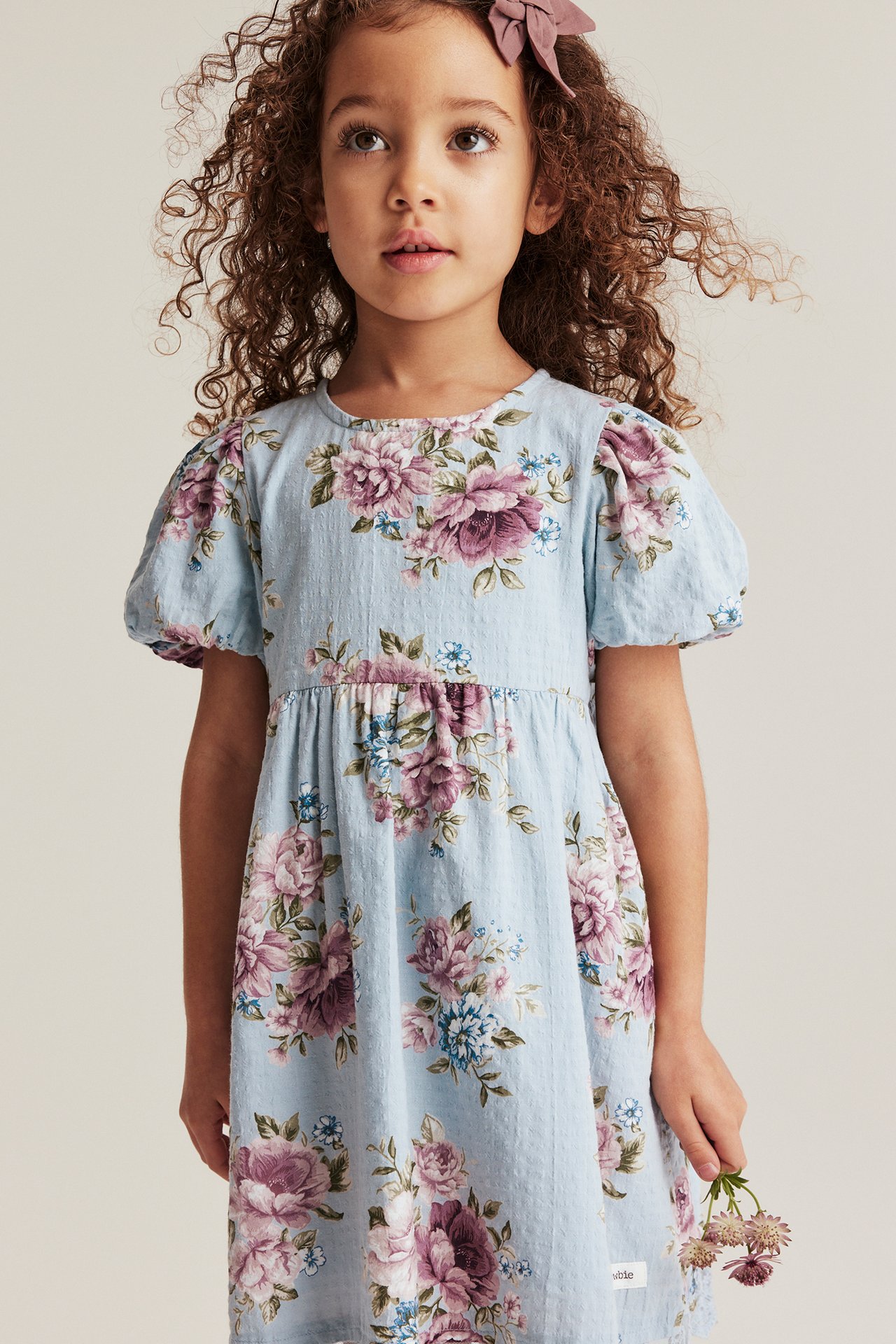 Vorderansicht eines Mädchens in hellblauem geblümtem Baumwollkleid mit Puffärmeln, hält Blumen.