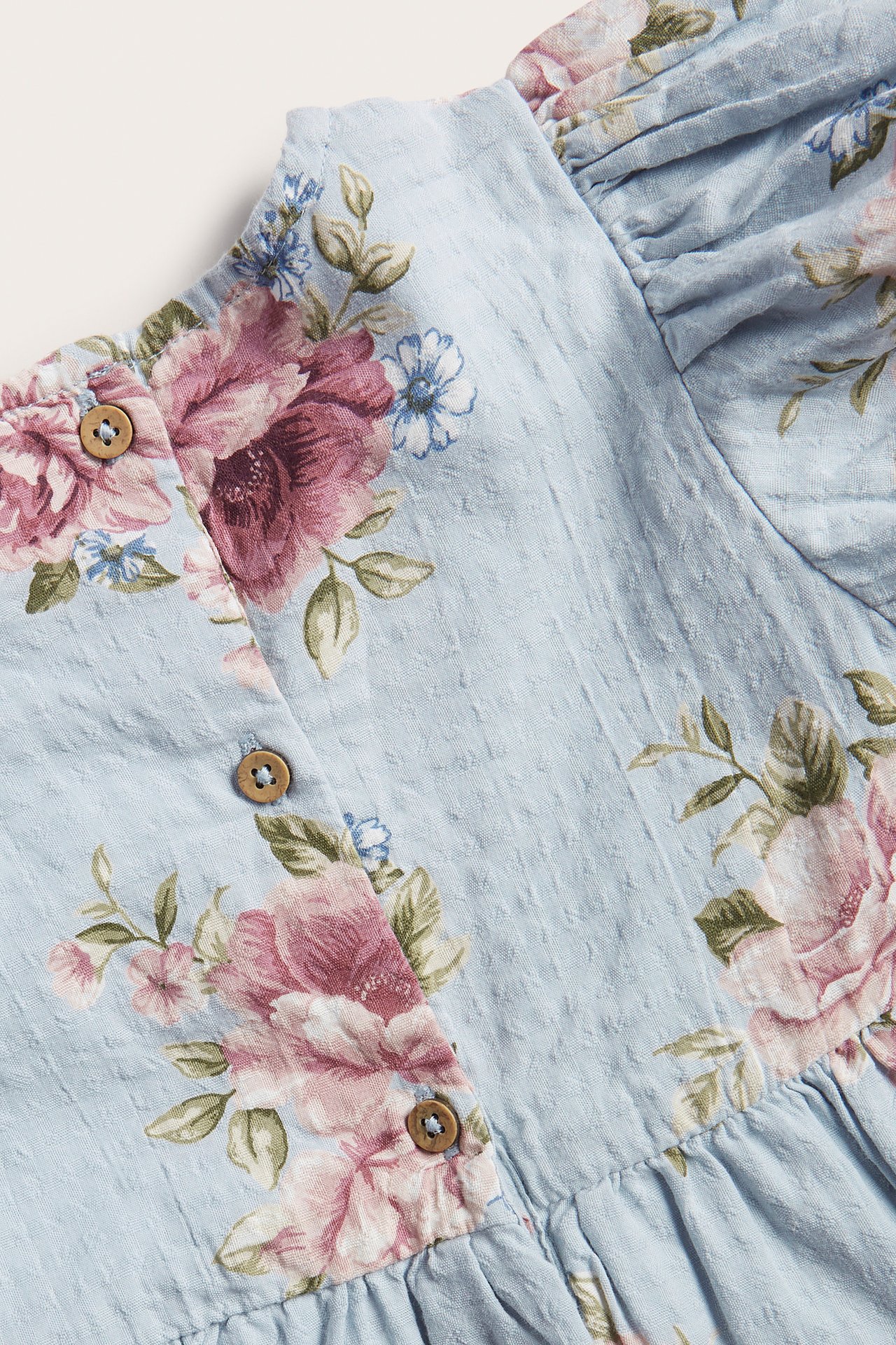 Nahaufnahme eines hellblauen gewebten Baumwollkleides mit rosa Blumenmuster und sichtbaren Holzknöpfen.