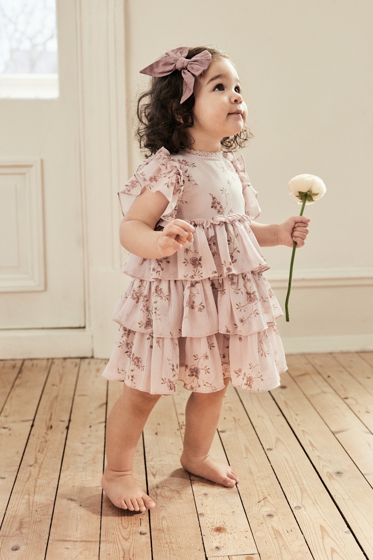 Vorderansicht eines Babys in einem rosa geblümten Chiffonkleid mit Rüschenärmeln, mit rosa Schleife, hält eine Blume.