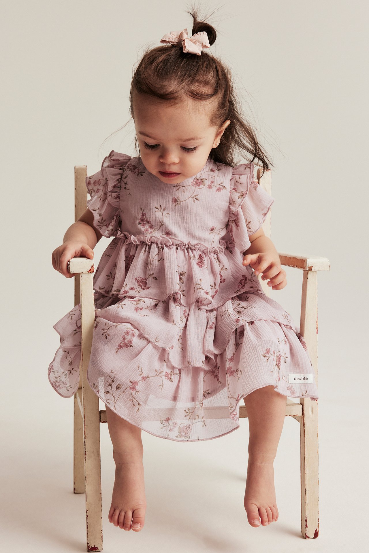 Vorderansicht eines Baby-Mädchens in einem rosa geblümten Chiffonkleid mit Rüschenärmeln und rosa Haarschleife.