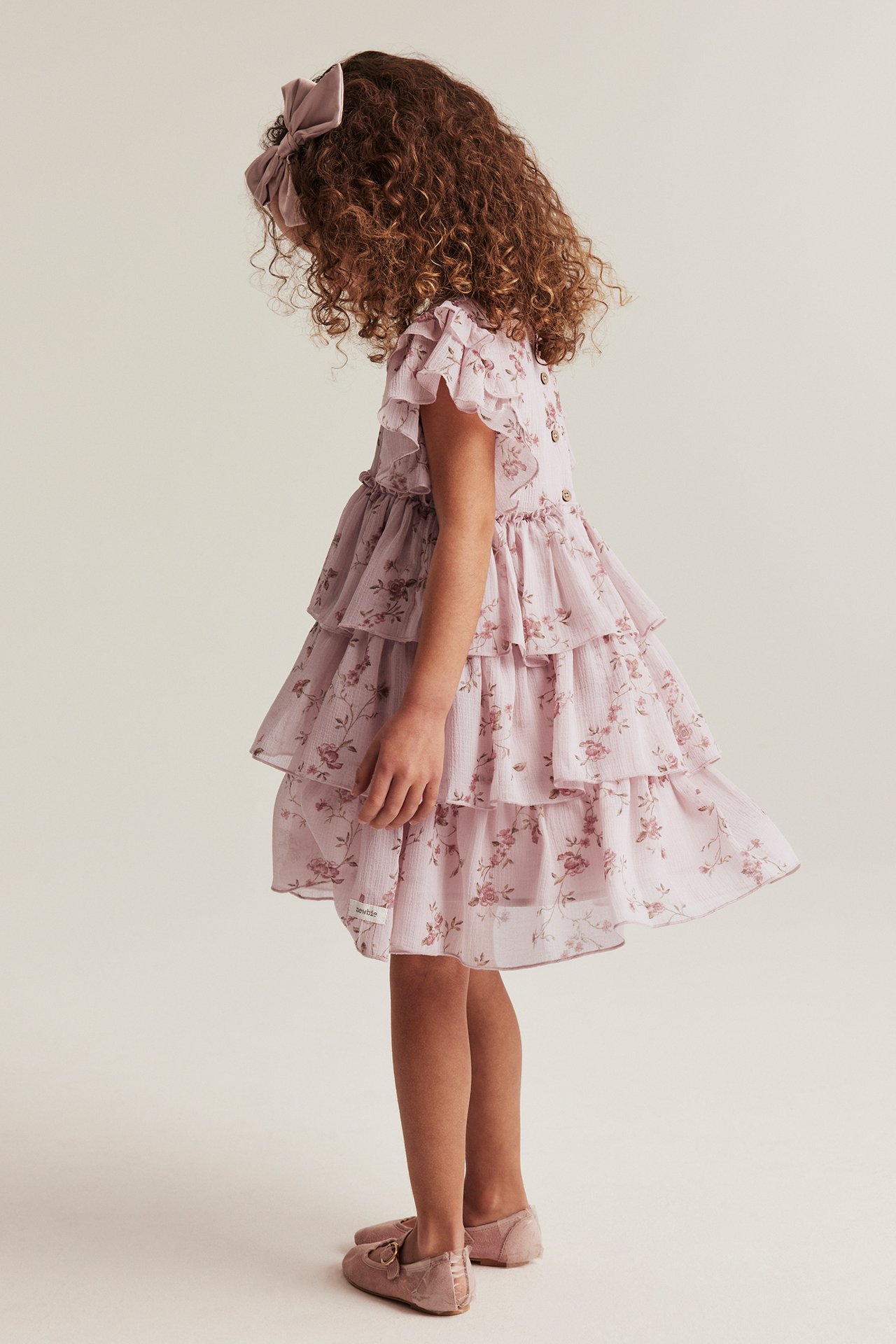 Side view of a kid wearing a pink floral tiered chiffon dress, a pink bow, and pink flat shoes.