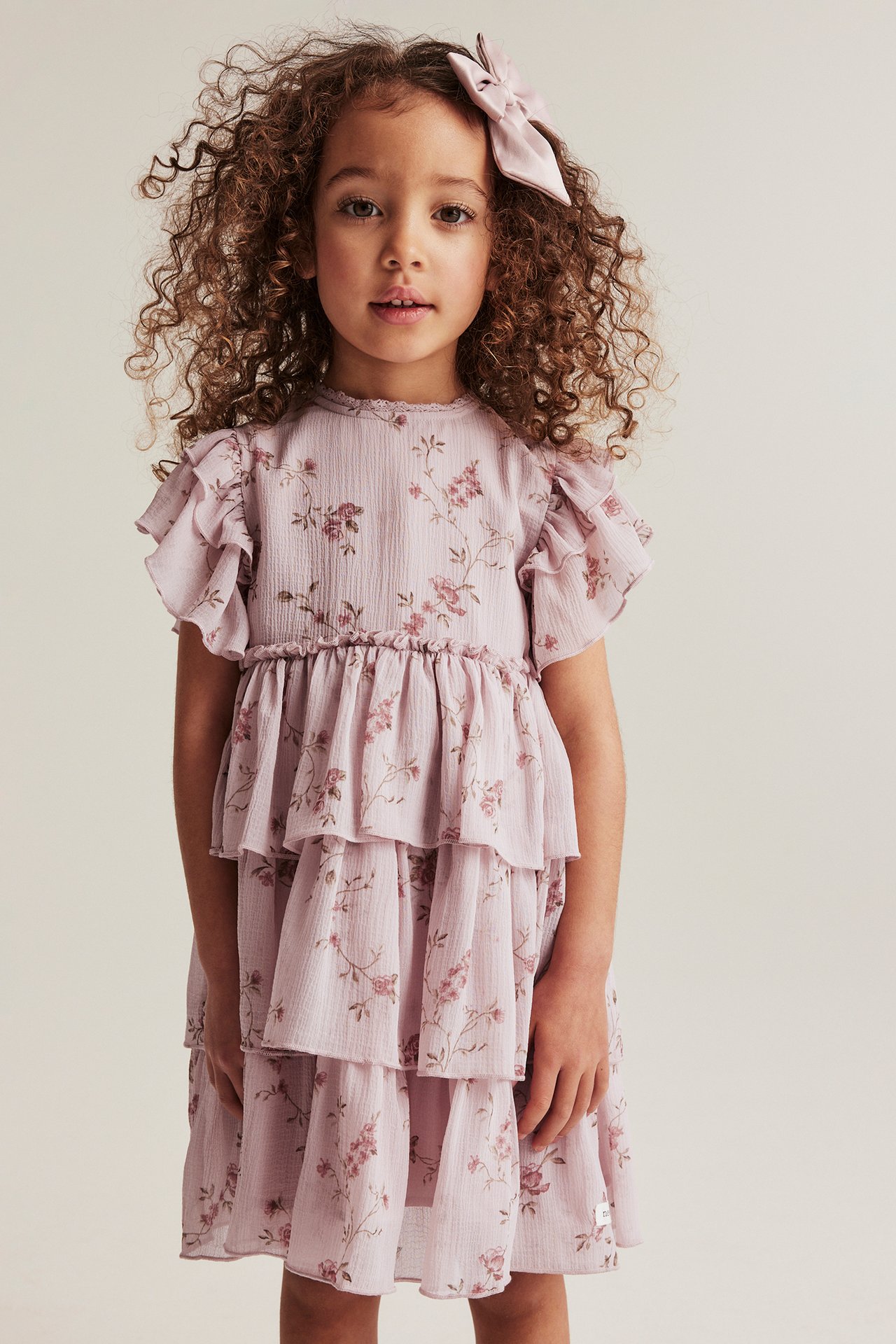 Front view of a kid wearing a pink floral tiered chiffon dress with ruffle sleeves and a pink hair bow.