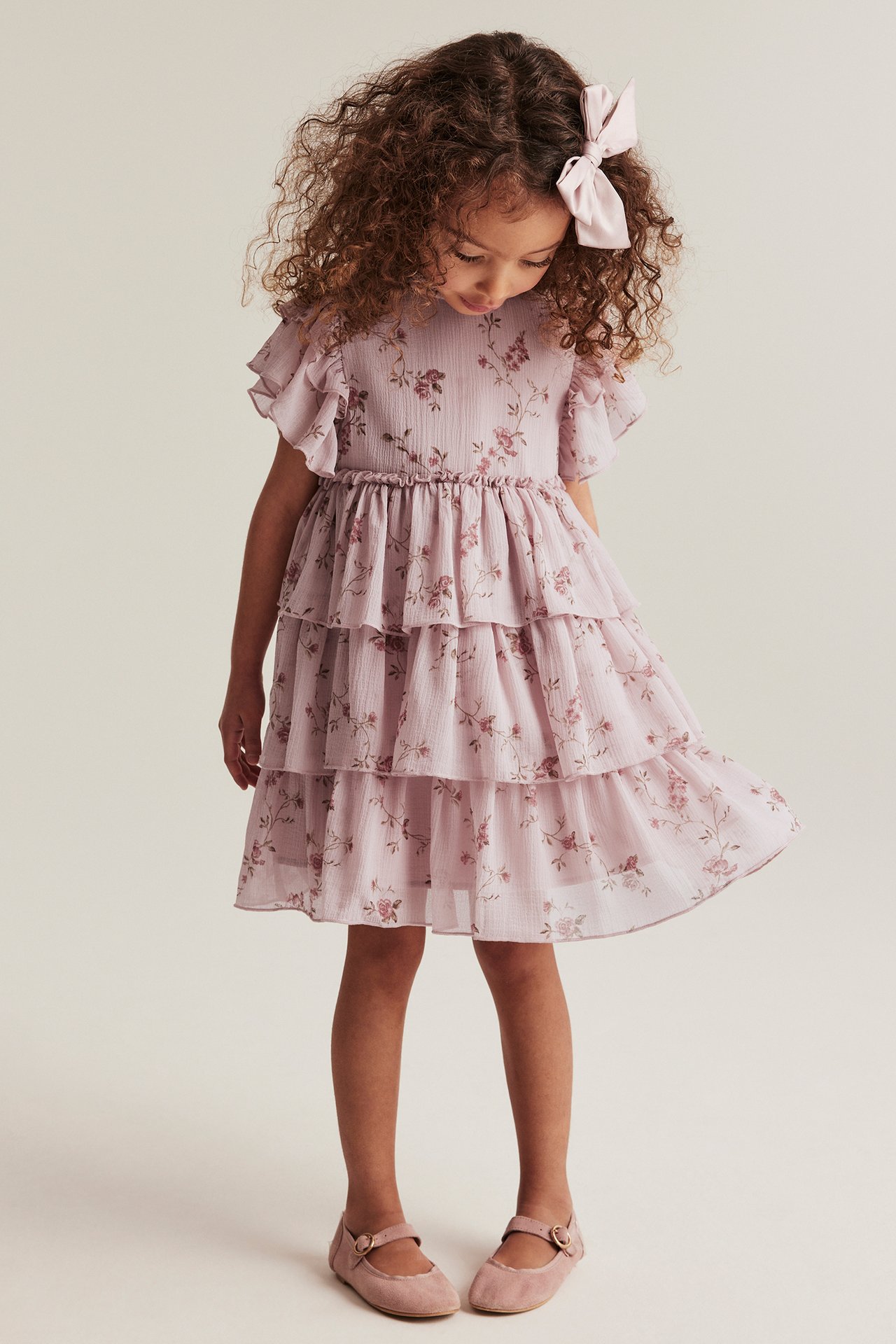 Front view of a kid wearing a pink floral tiered chiffon dress, pink bow, and pink Mary Jane shoes.