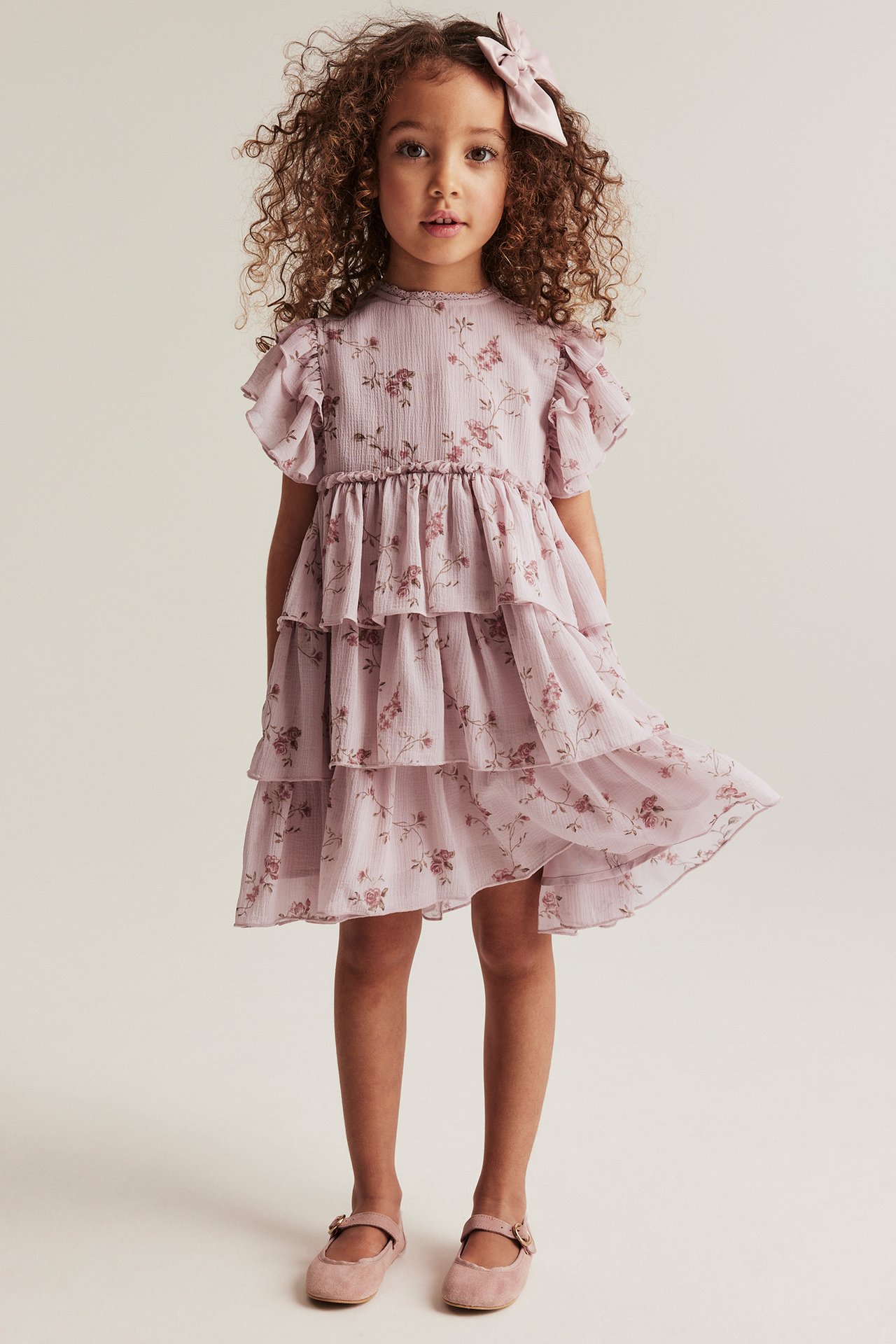 Front view of a kid wearing a pink floral ruffled chiffon dress, a pink bow, and pink Mary Jane shoes.