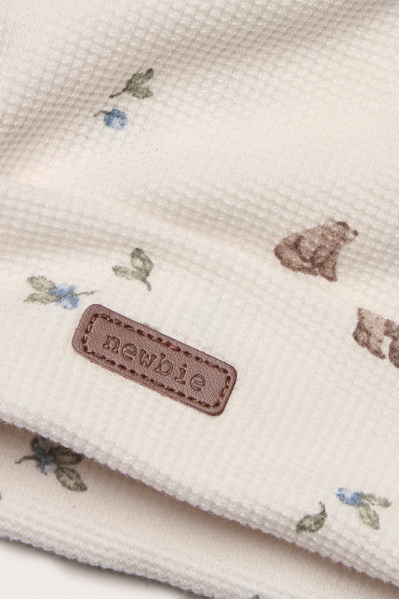 Close-up of an off-white waffle knit beanie with bear and foliage pattern and a brown 'newbie' label.