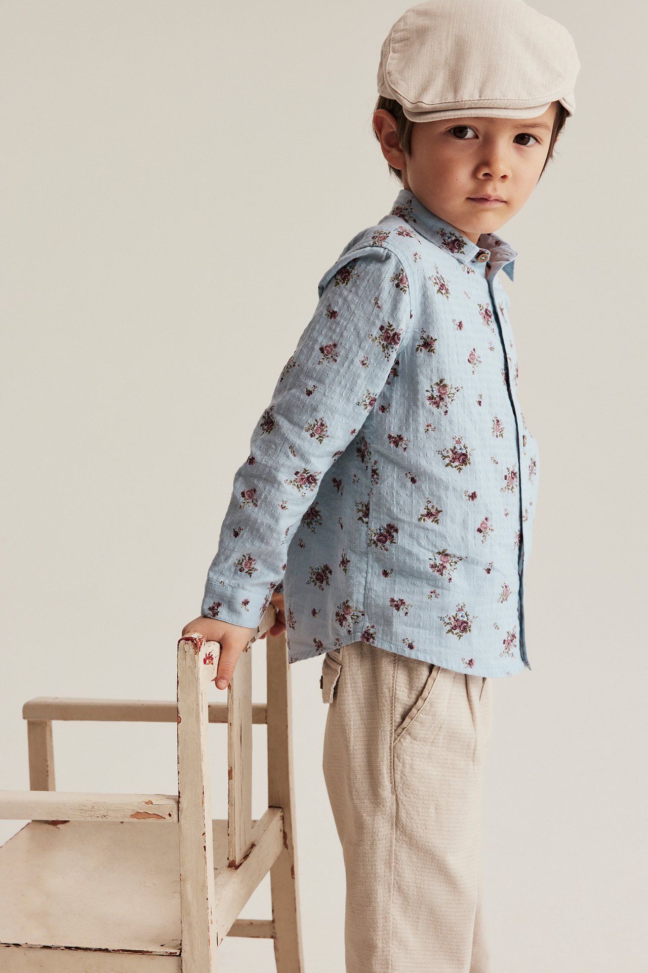 Front view of a kid wearing a light blue cotton floral shirt, beige trousers, and a beige flat cap.