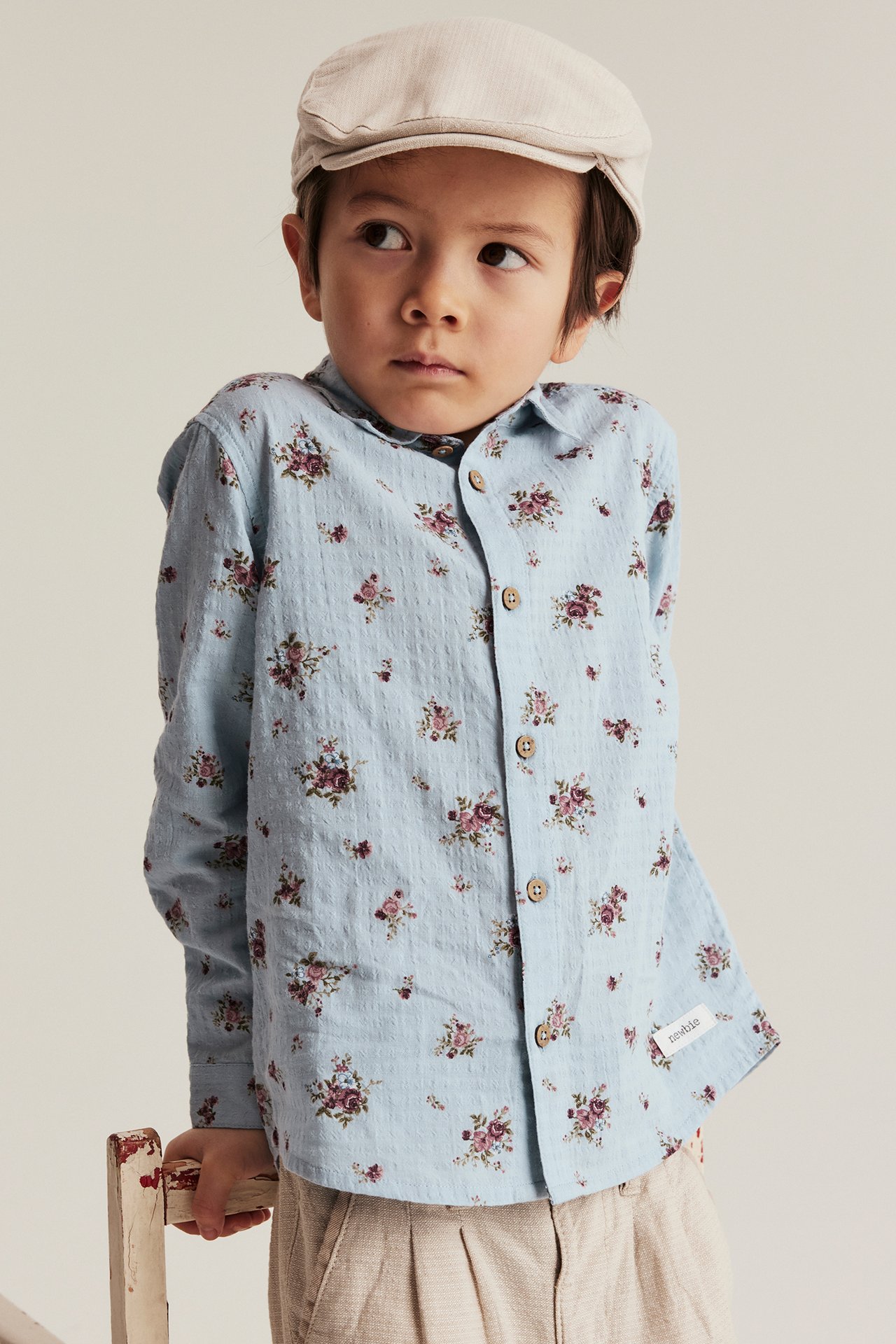 Front view of a kid wearing a light blue organic cotton shirt with small floral print, beige cap, and beige trousers.