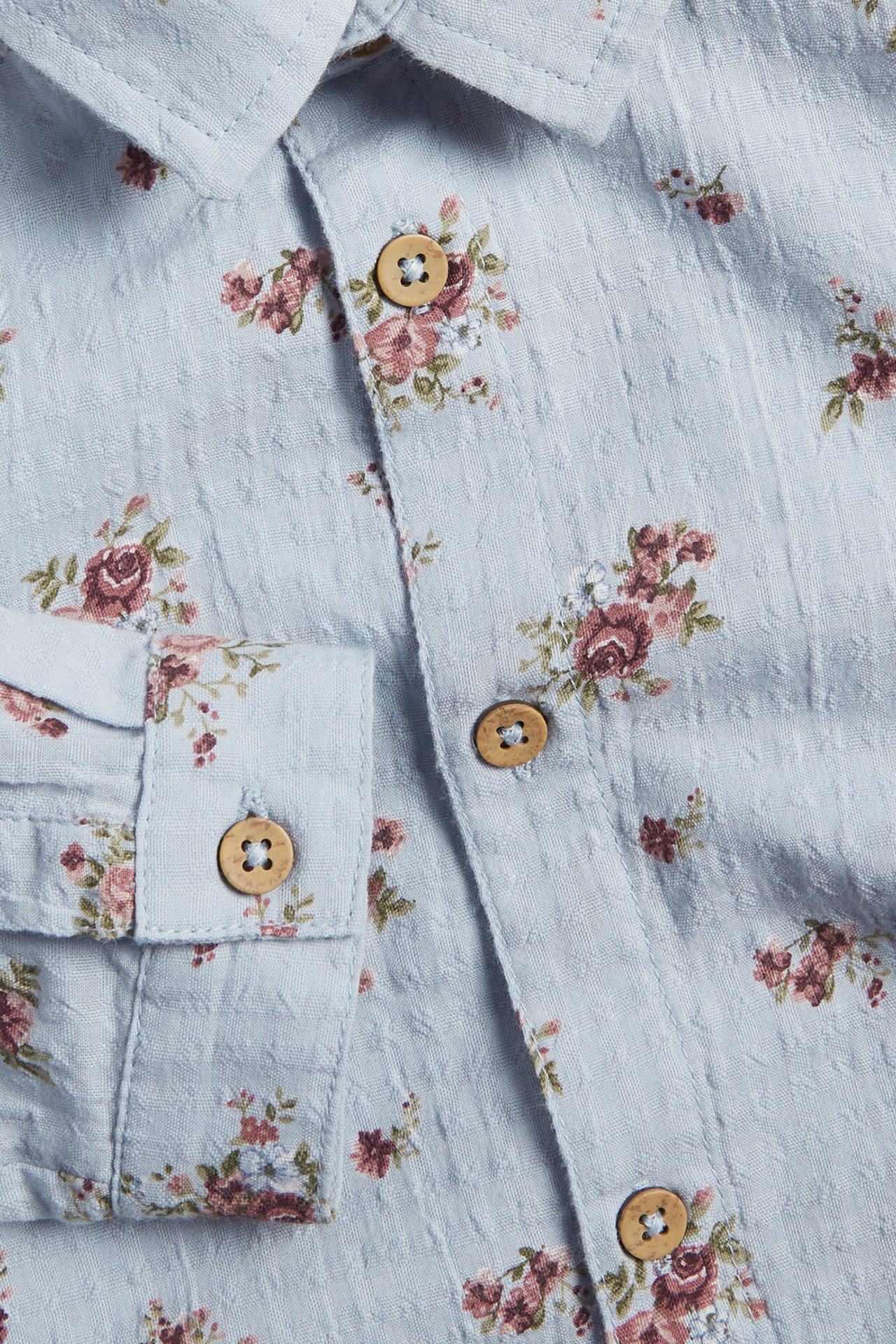 Close-up of a light blue woven cotton shirt with a pink floral pattern and wooden buttons.