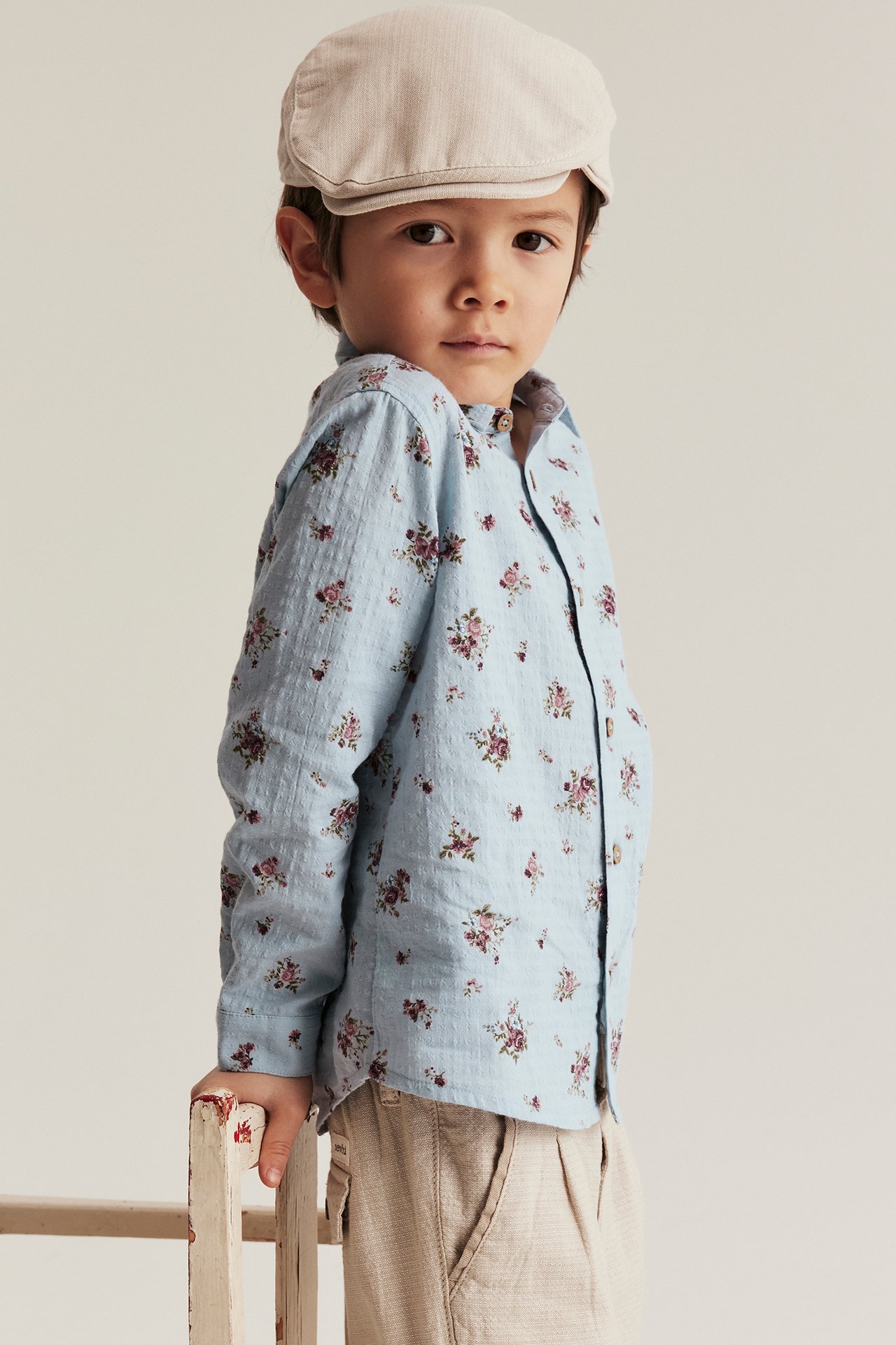 Front view of a kid wearing a light blue floral cotton shirt, beige trousers, and a beige flat cap.