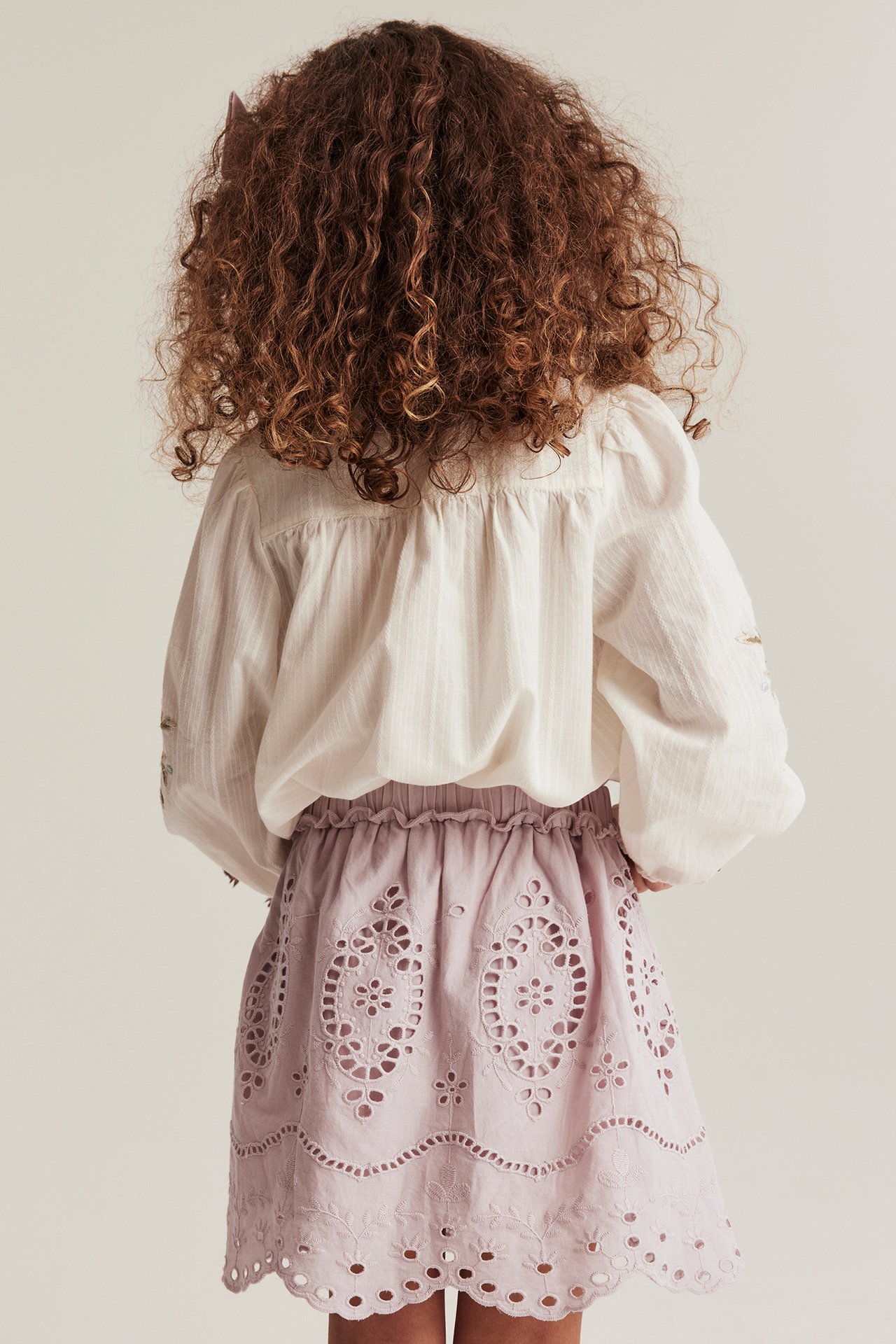 Back view of a kid in a light purple embroidered skirt with eyelet pattern and a white long-sleeved blouse.
