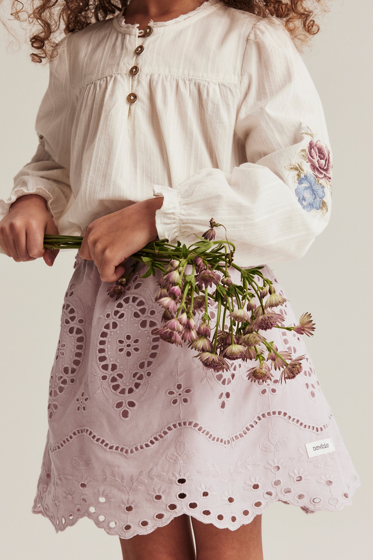 Front view: Kid in pink embroidered eyelet skirt and off-white blouse with floral sleeves, holding flowers.