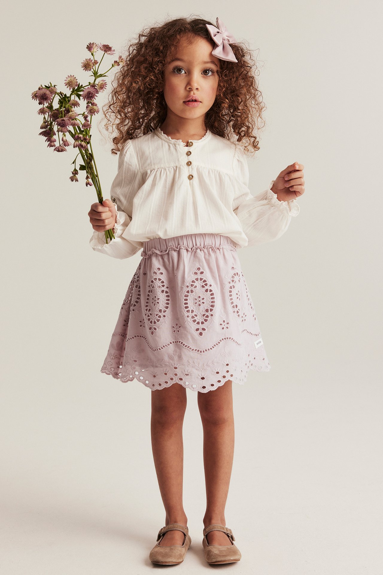 Front view: Kid in pink embroidered A-line skirt with elastic waist, white blouse, beige shoes, holding flowers.