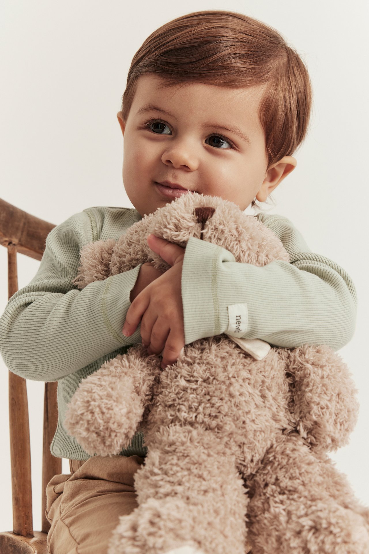 Vorderansicht eines Babys in grünem geripptem Langarmbody, brauner Hose, das einen beigen Teddybär hält.