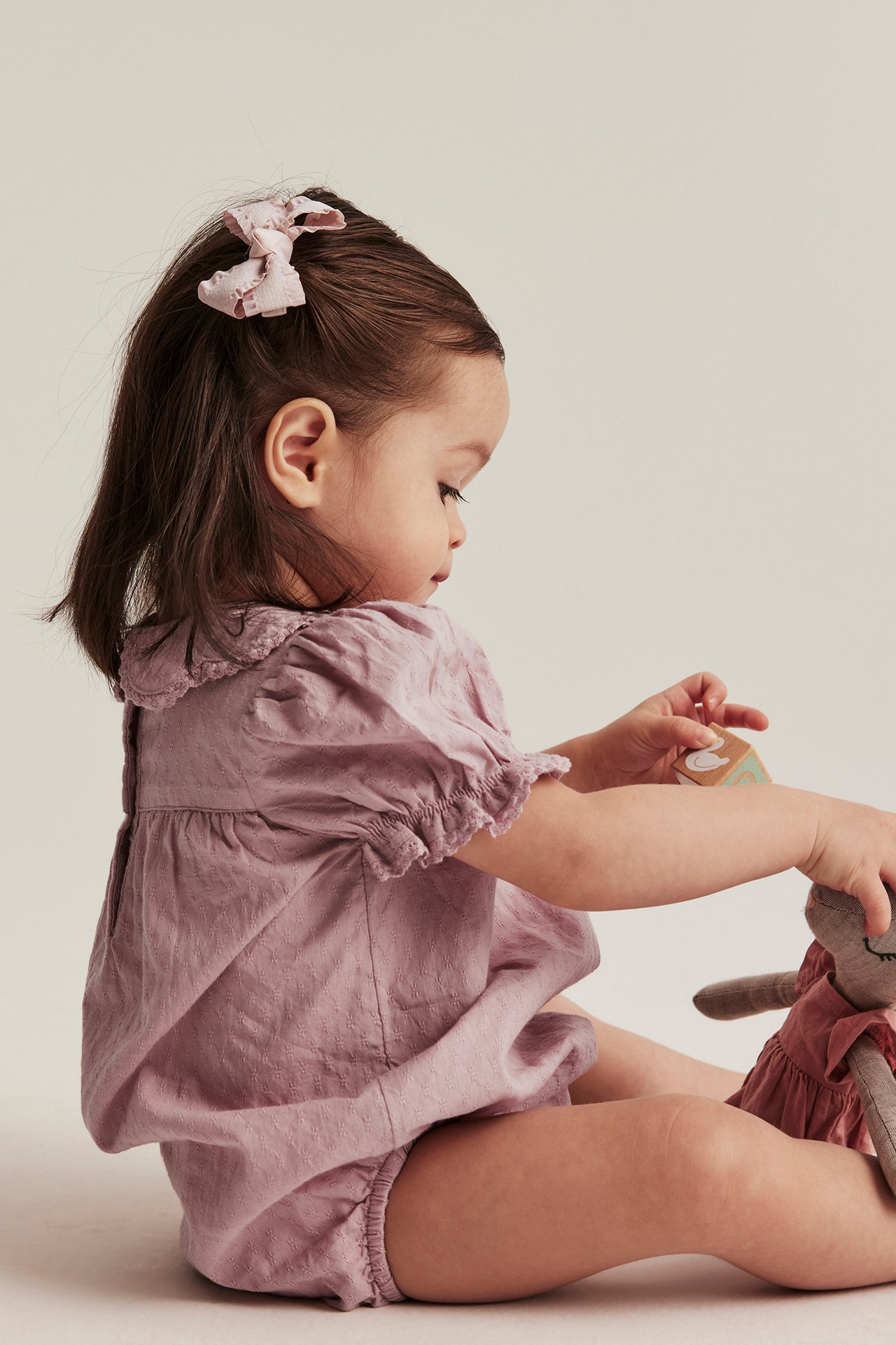Side view of baby in pink textured cotton body with lace collar, pink bow, holding doll and block.