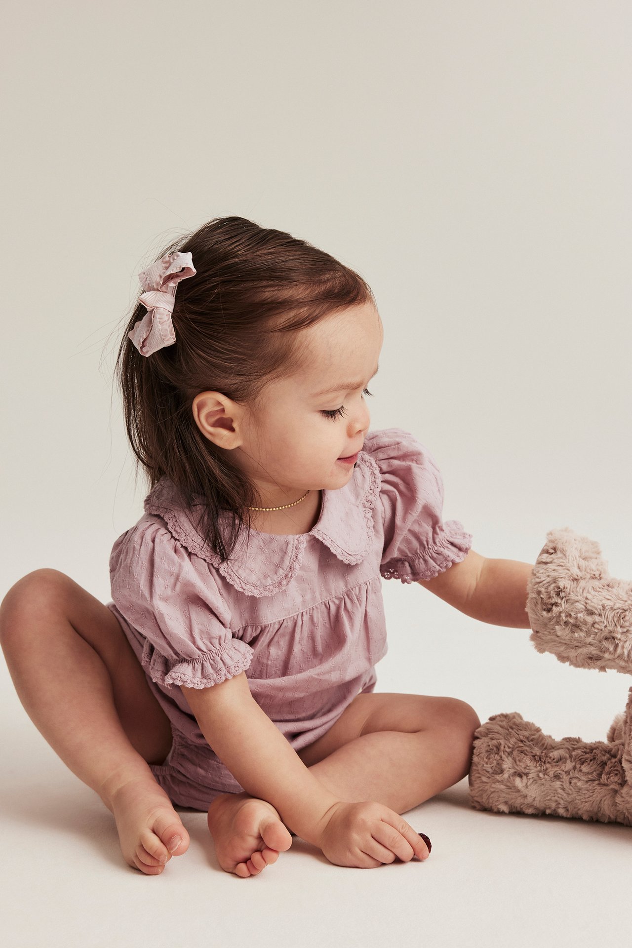 Side view: baby in pink woven body with lace collar, puff sleeves, pink bow, and plush toy.