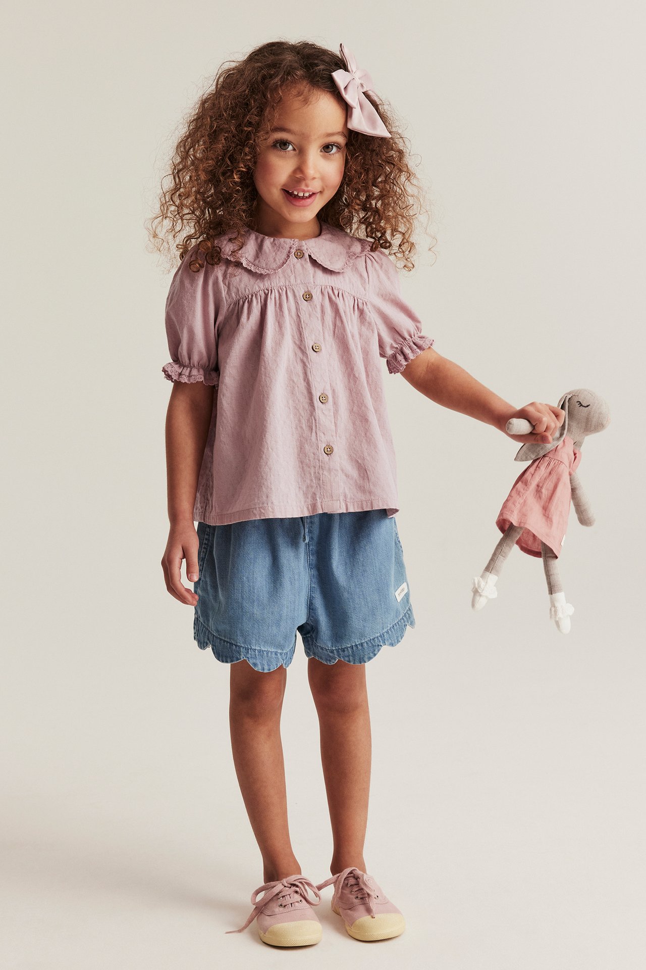 Kind in rosa strukturierter Bluse, blauen Shorts, rosa Schuhen, rosa Schleife, hält eine Hasenpuppe.