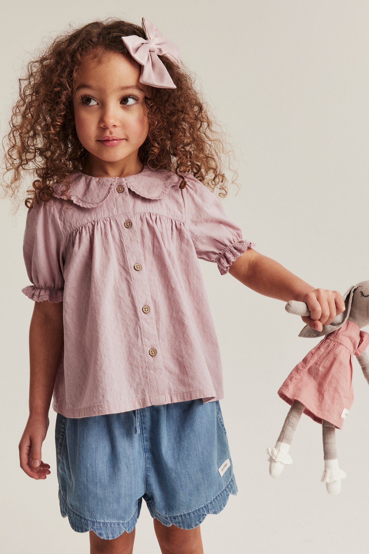Vorderansicht: Kind in rosa Baumwollbluse mit Spitzenkragen, blauen Shorts, rosa Schleife, hält eine Hasenpuppe.