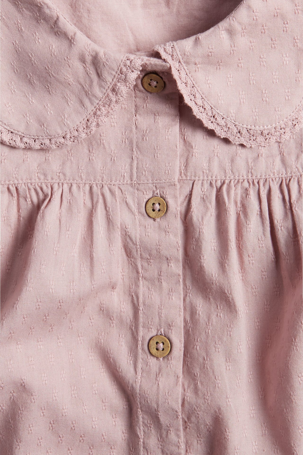 Nahaufnahme von vorne einer rosa strukturierten Baumwollbluse mit Spitzenkragen und Holzknöpfen.