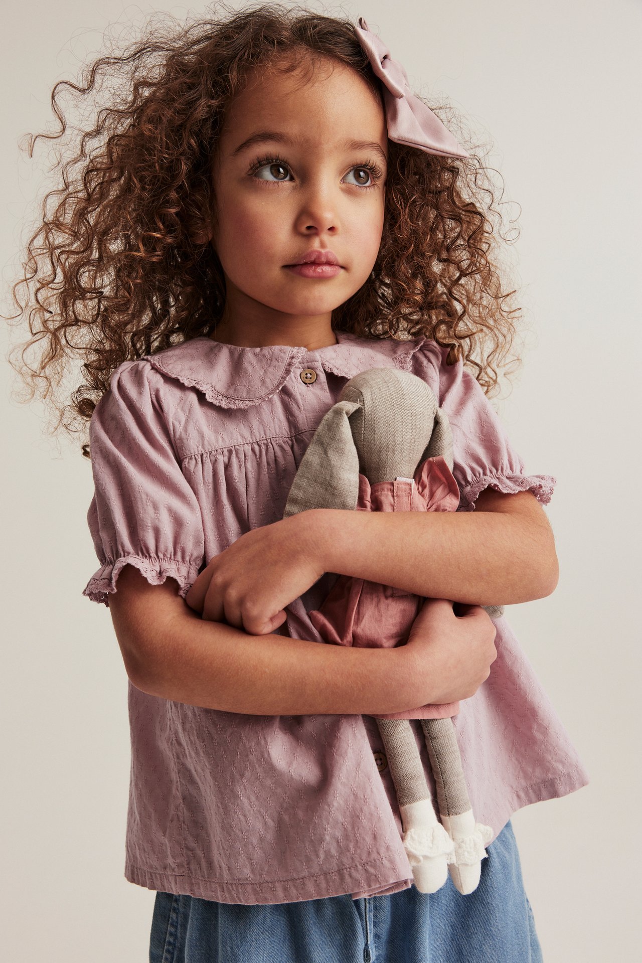 Vorderansicht: Mädchen in rosa strukturierter Bluse mit Spitzenkragen, blauen Shorts, rosa Schleife, hält eine Puppe.