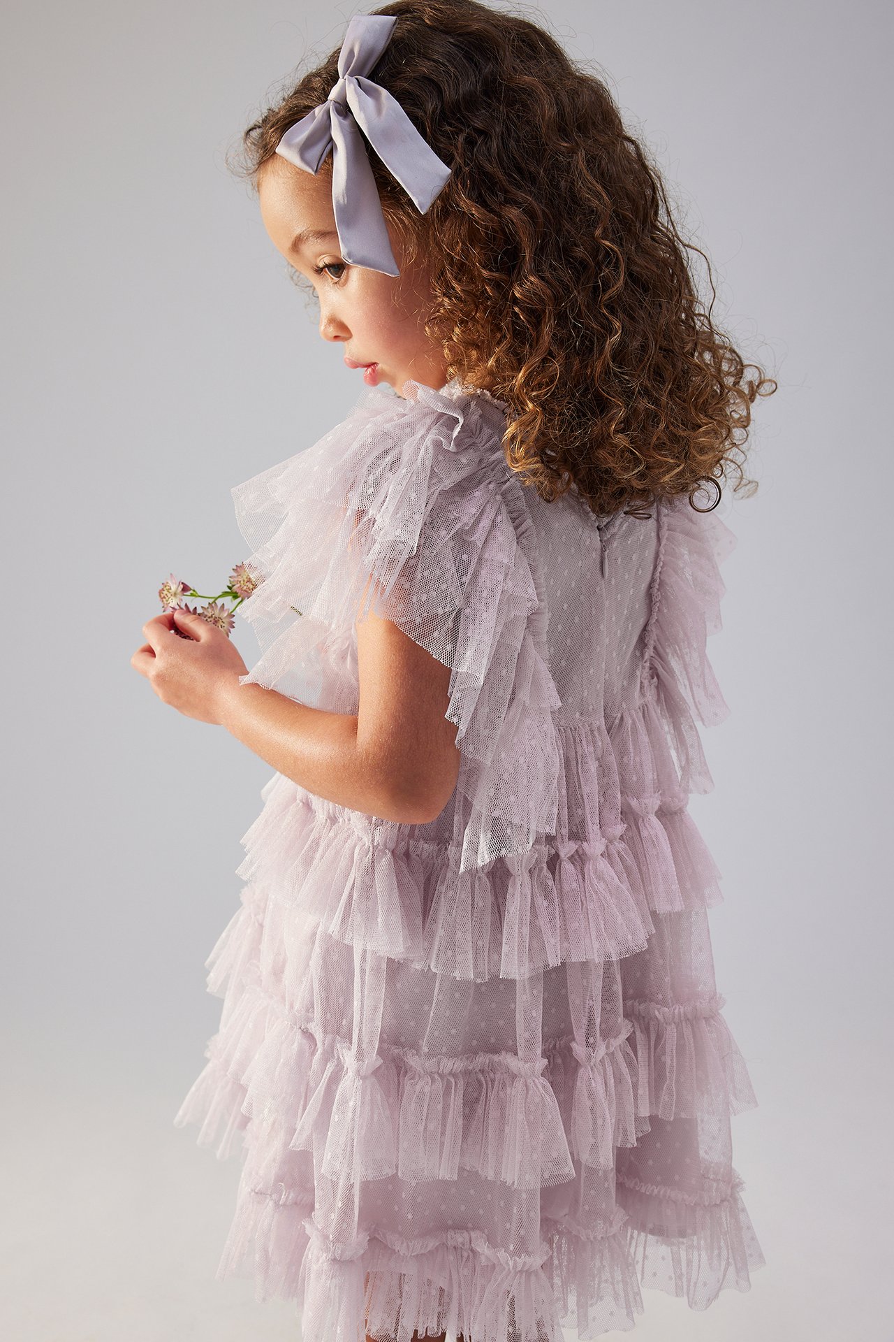 Side view of a kid in a light lilac dotted mesh dress with ruffles, wearing a grey bow, holding flowers.