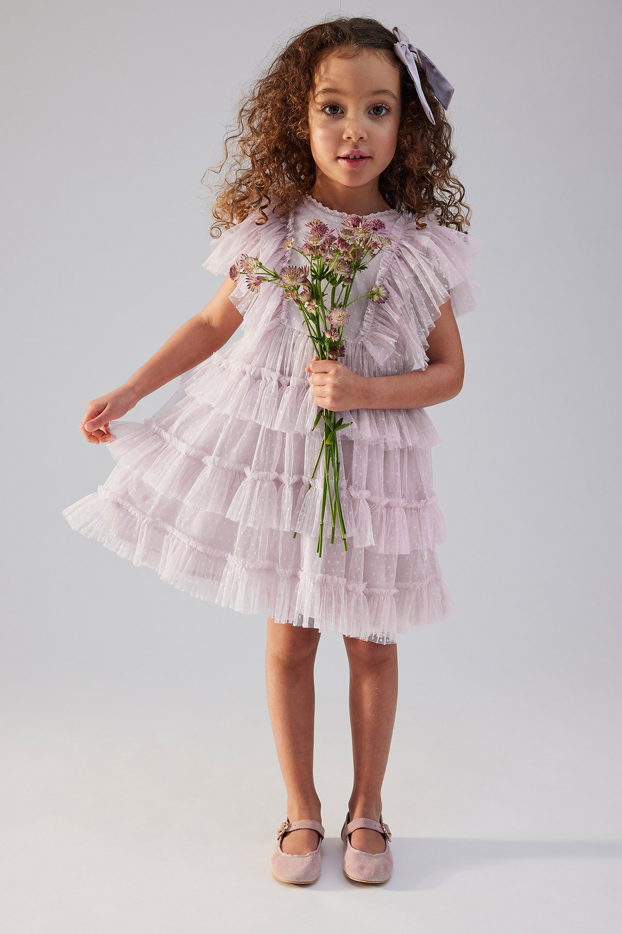 Front view of a kid in a light lilac tiered mesh dress with polka dots, holding flowers, wearing pink shoes.
