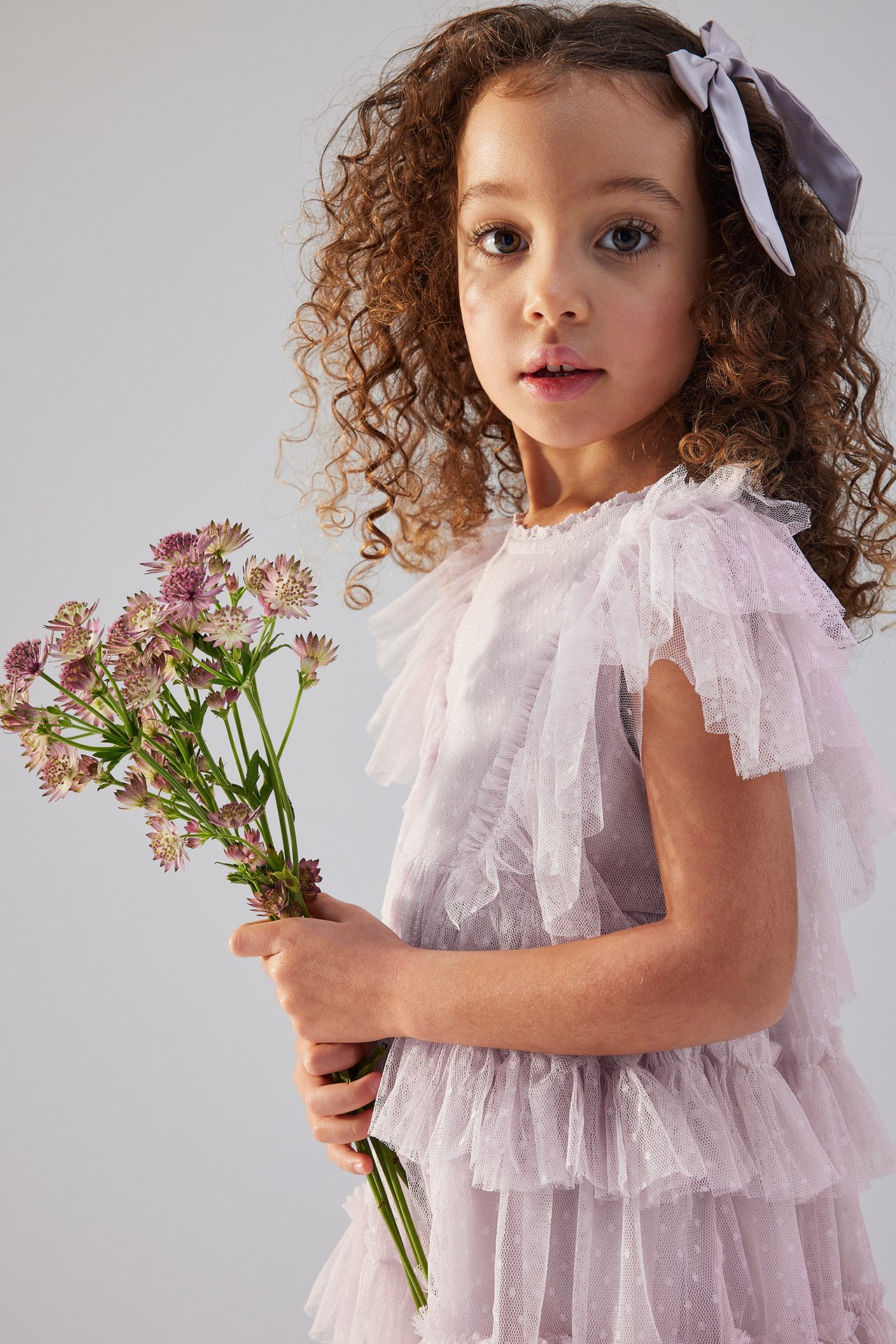 Front view of a kid in a light lilac ruffled mesh dress with polka dots, a grey hair bow, holding flowers.