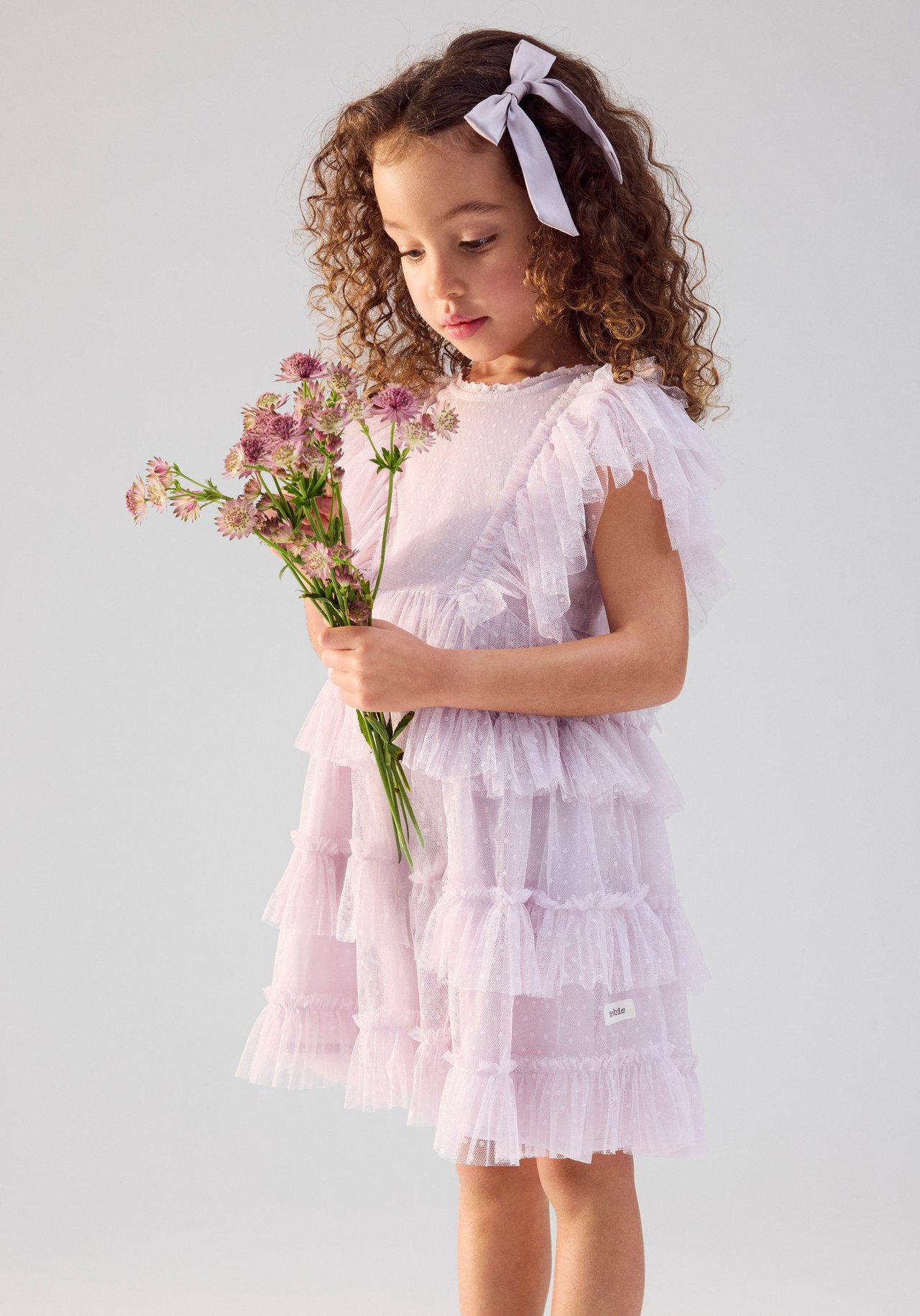 Front view of a kid wearing a light lilac dotted mesh dress with ruffles, holding pink flowers, and a lilac hair bow.