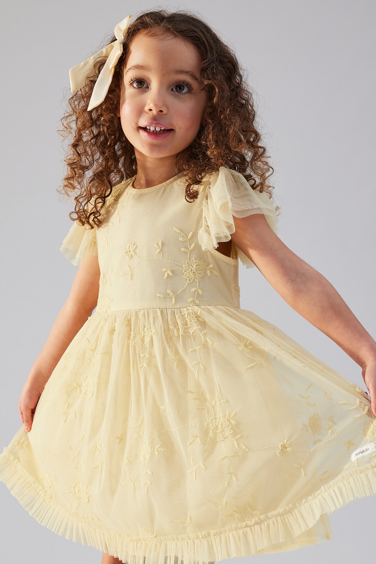 Front view of a kid wearing a light yellow embroidered dress with ruffled sleeves and a cream hair bow.