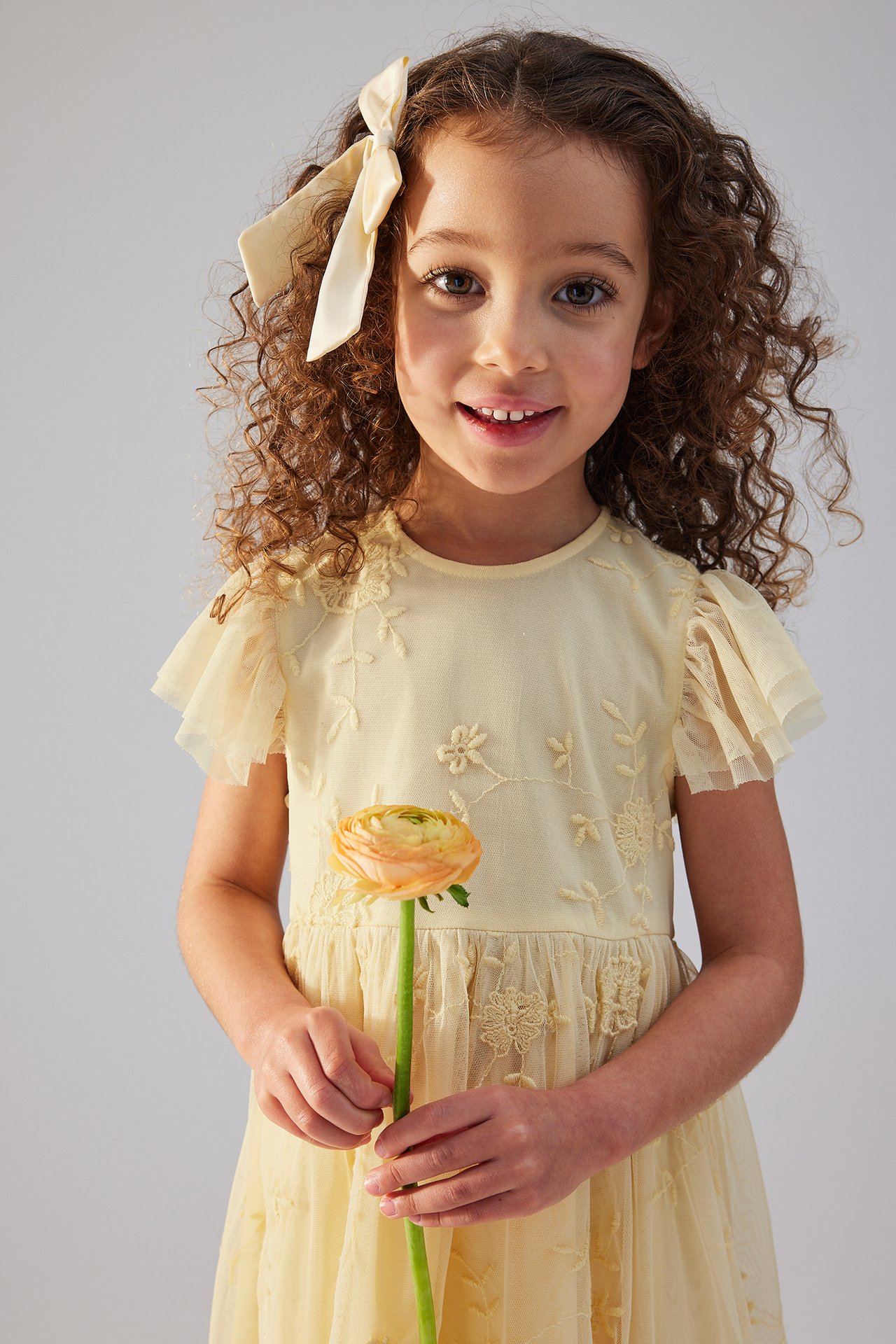 Front view of a kid wearing a yellow embroidered dress with frill sleeves, a cream hair bow, holding an orange ranunculus.