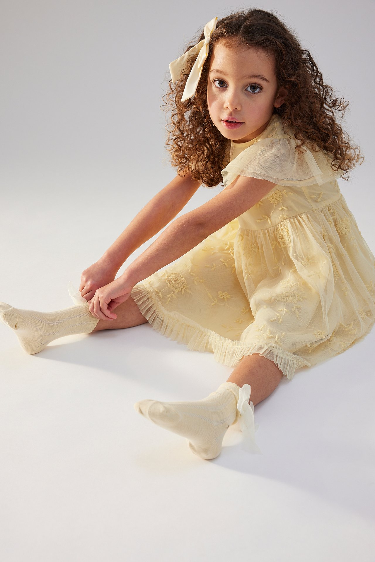 Front view of a kid wearing a yellow embroidered dress with frills, cream socks, and a yellow bow.