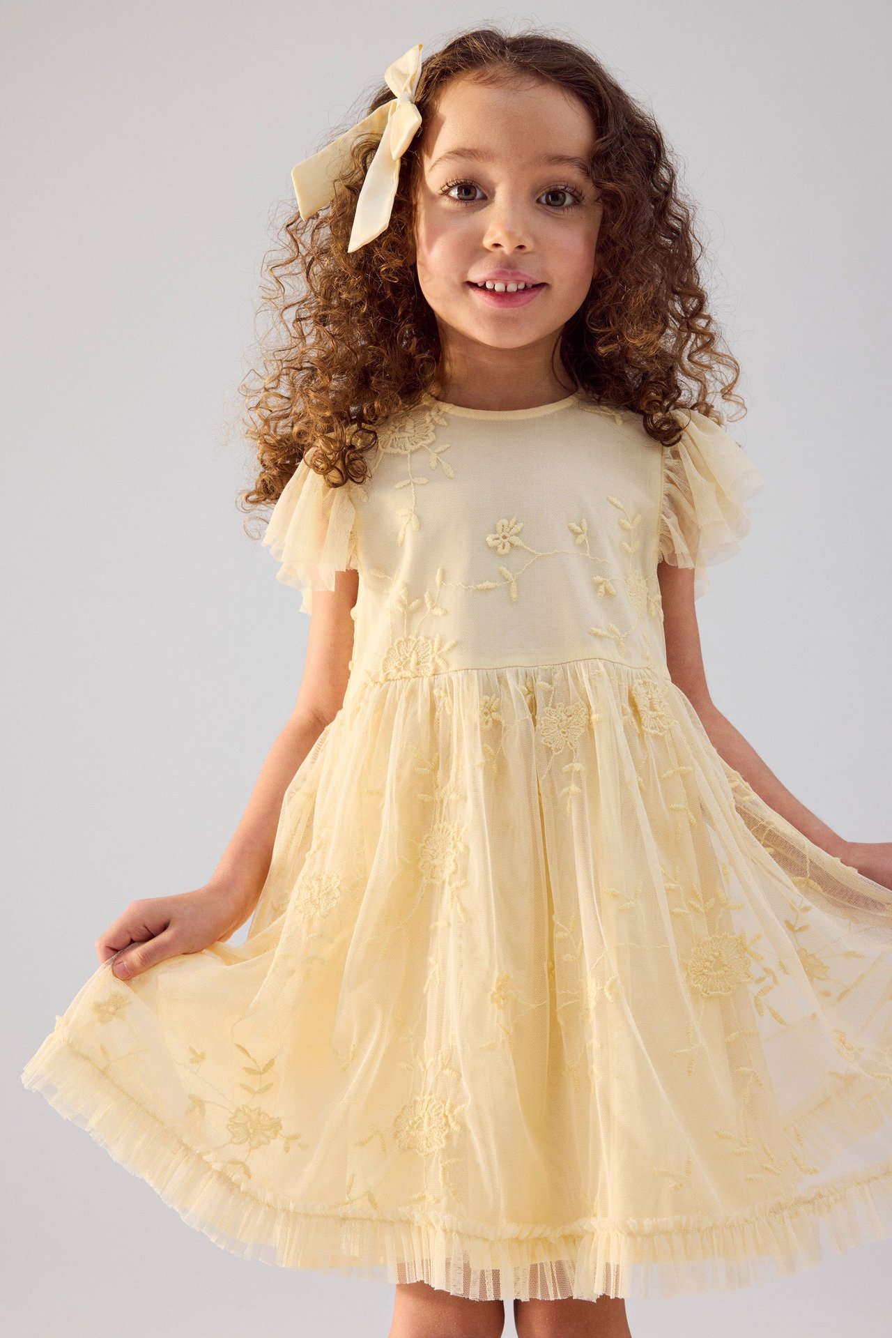 Vorderansicht eines Kindes in einem gelben bestickten Kleid mit Rüschenärmeln und passender Haarschleife.