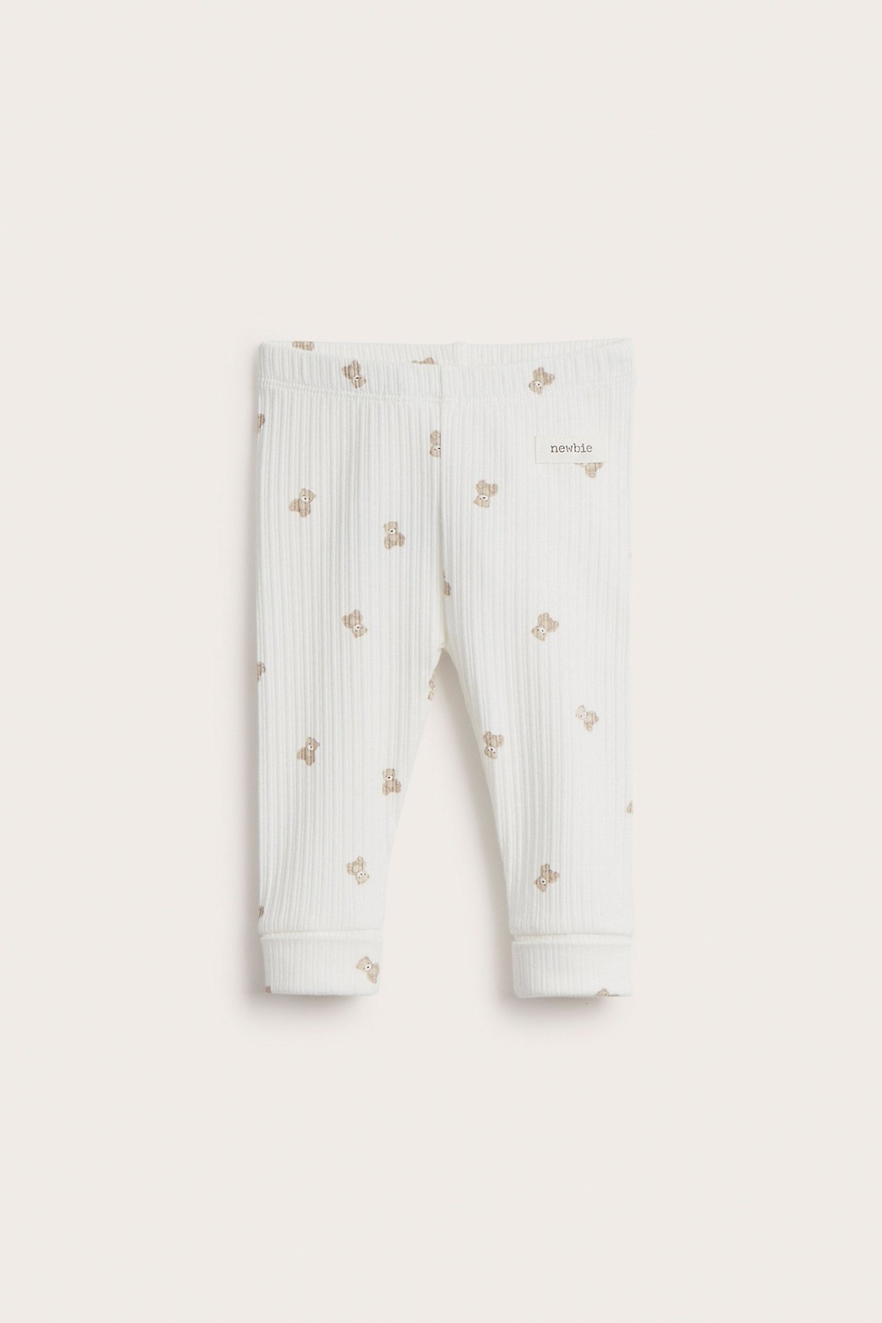 Front view of off-white ribbed baby leggings with a brown teddy bear pattern and a 'newbie' tag.