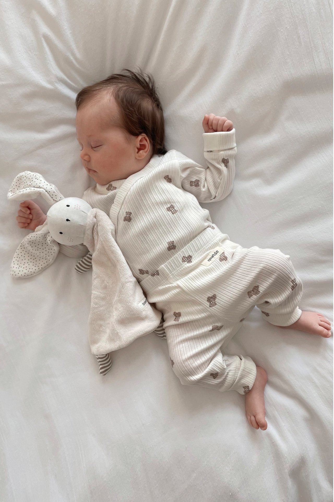 Top view: sleeping baby in off-white ribbed teddy bear patterned wrap bodysuit, next to a bunny comforter.